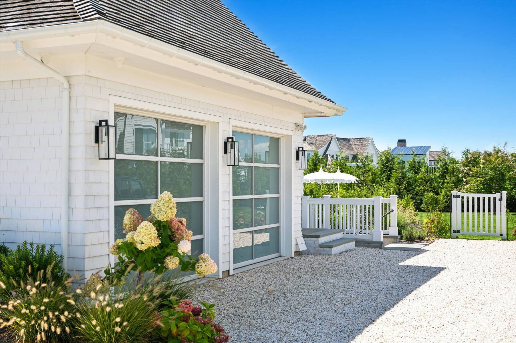 White shingle house exterior with large windows, gravel path, white-railed steps, and flower beds.