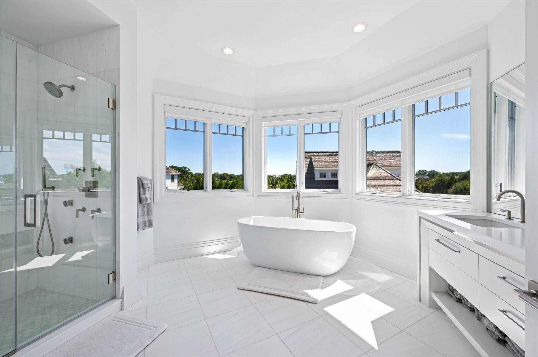 Bright white bathroom with a freestanding soaking tub, glass shower, and large windows overlooking green trees.
