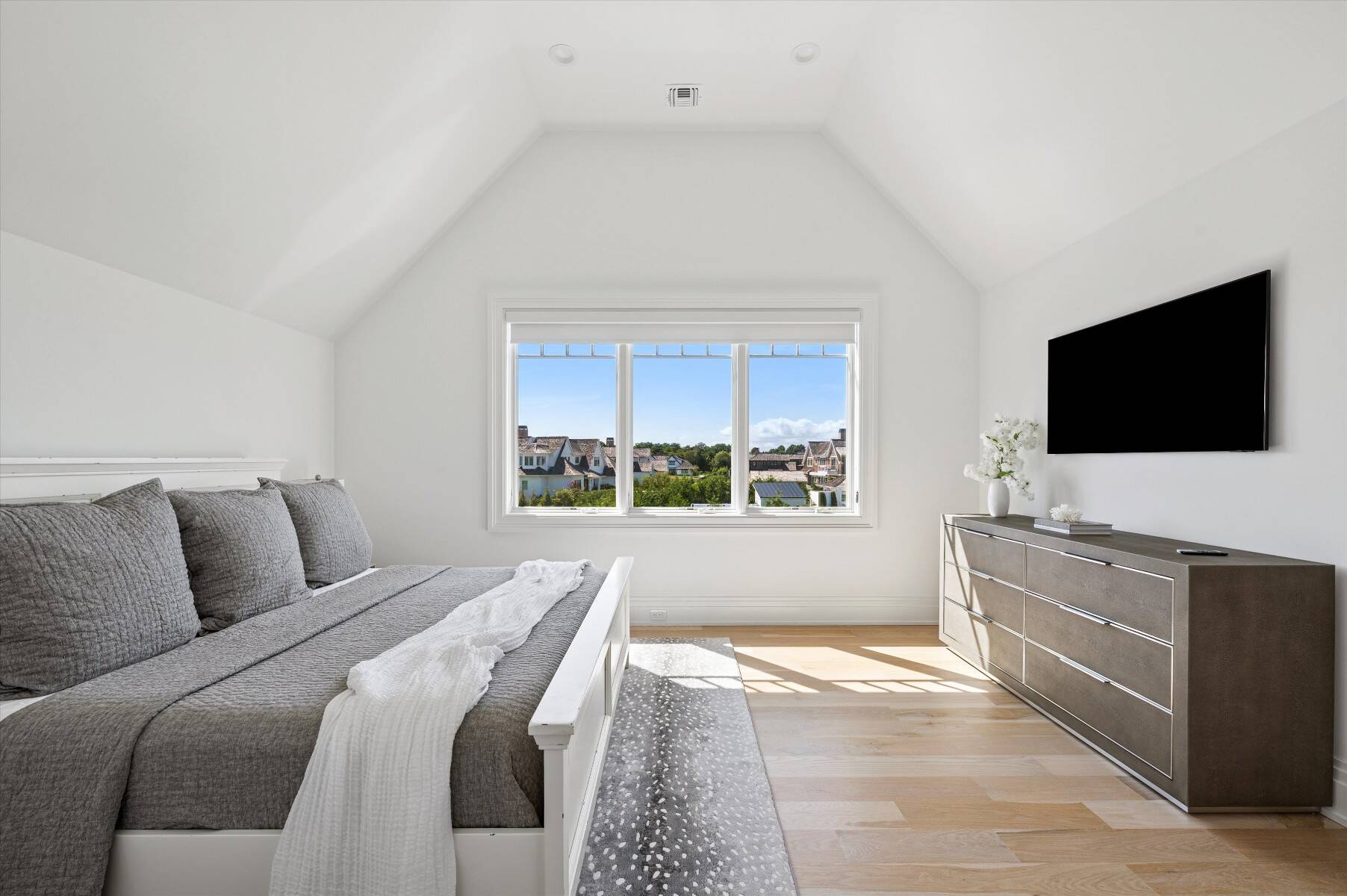 Bright bedroom with gray bedding, vaulted ceiling, and a window overlooking neighboring homes.