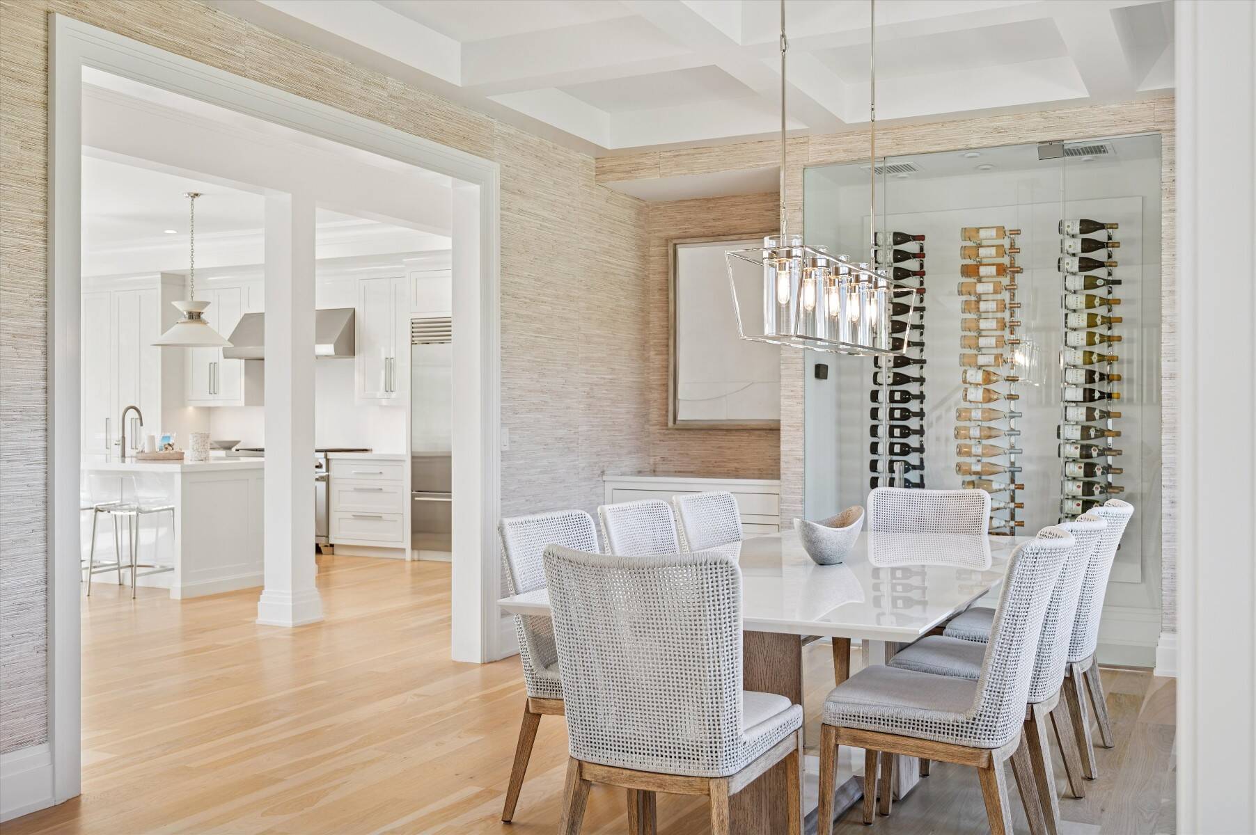 Dining room with woven chairs, a glass wine display, and an adjoining white kitchen.