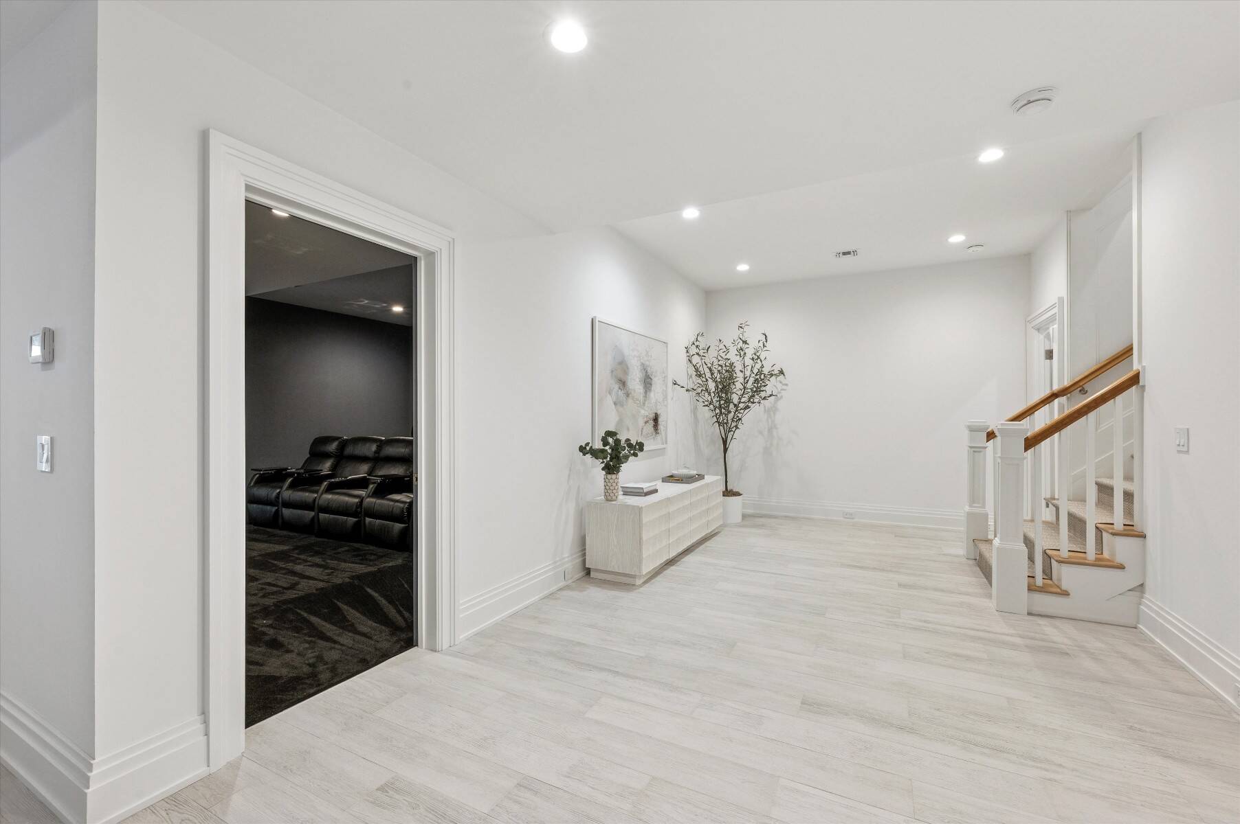 Bright white hallway with a staircase and console table beside an open doorway to a dark media room.