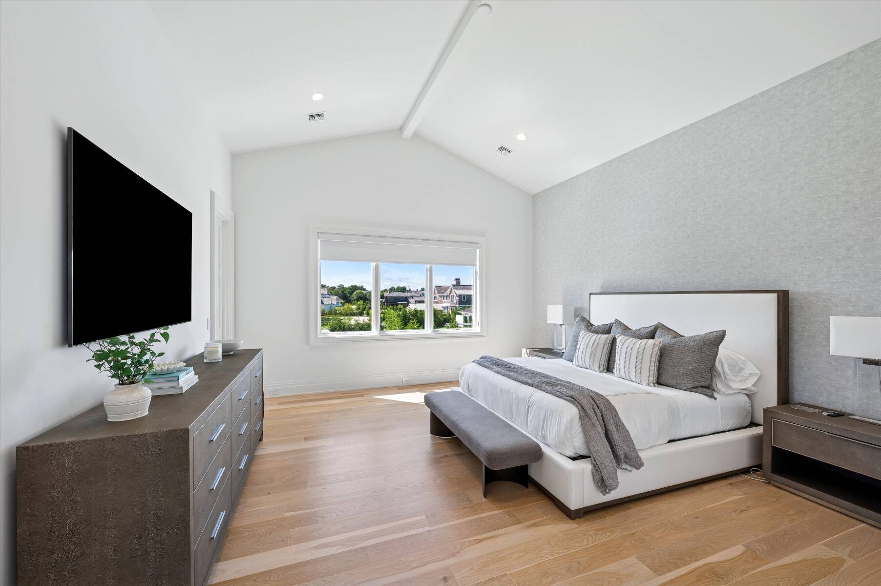 Furnished bedroom with a white bed, gray accent wall, and window overlooking greenery.