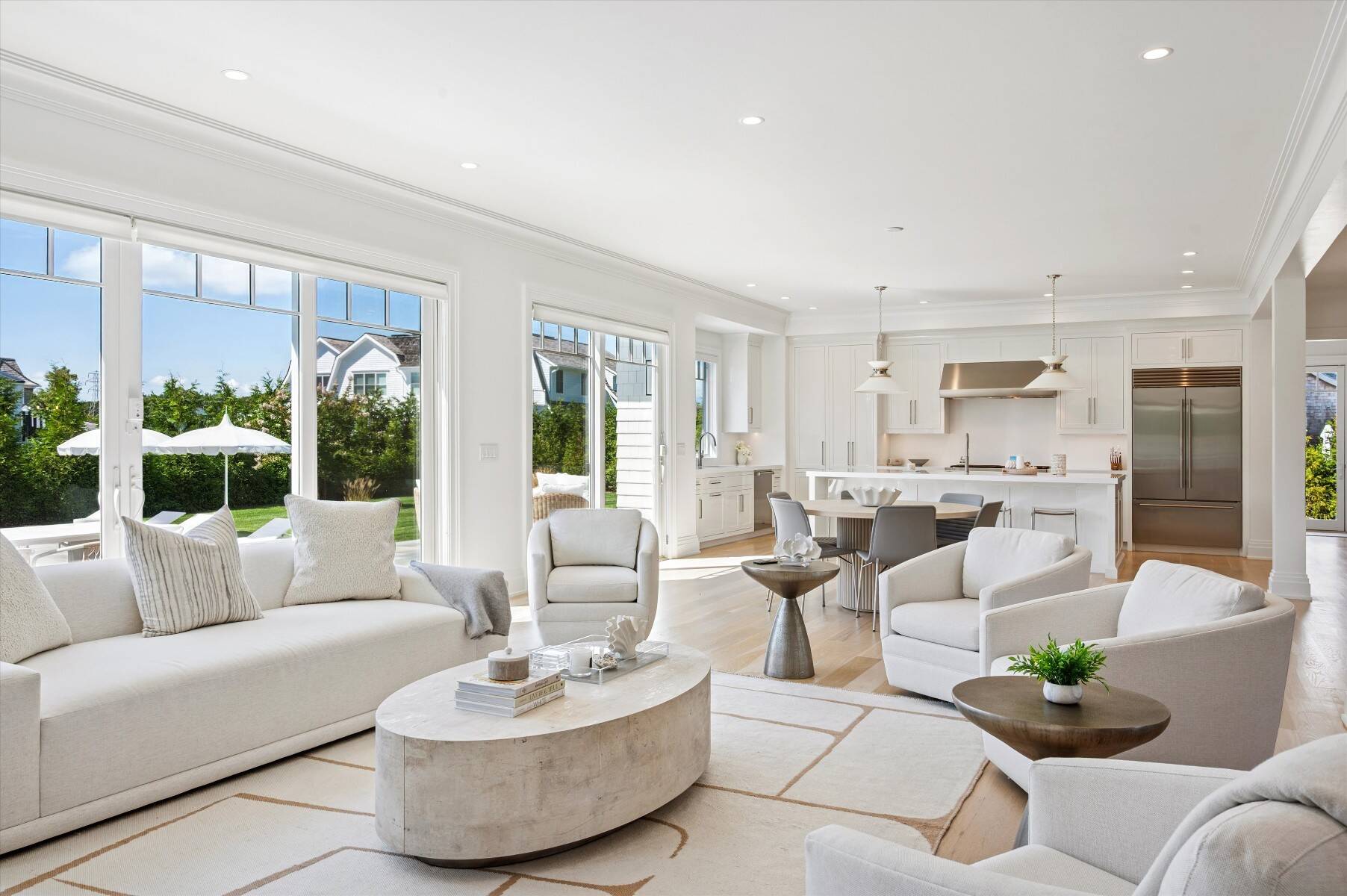 Bright open-plan living room with white sofas, large windows, and an adjoining kitchen.