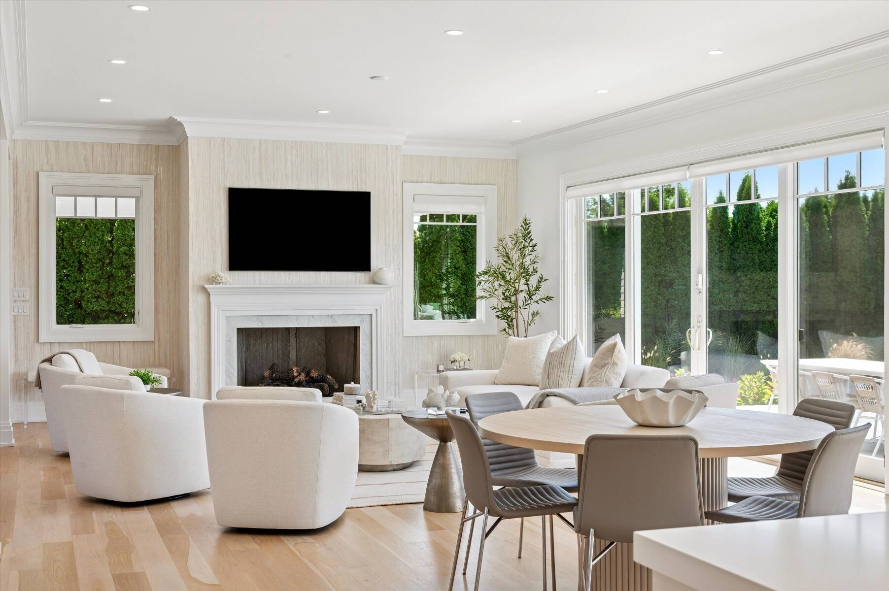 Living and dining room with a white fireplace, cream seating, and large windows overlooking hedges.
