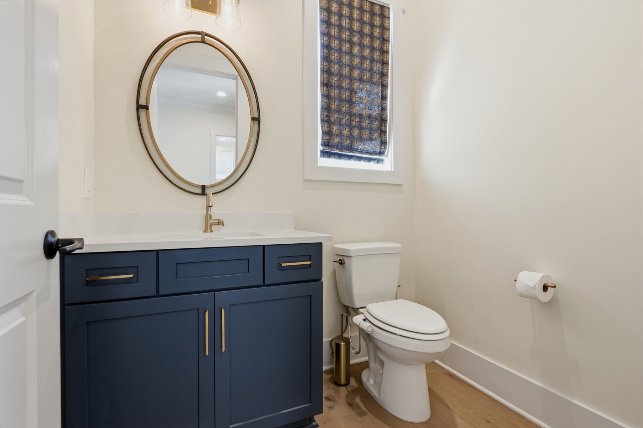 Bathroom with navy vanity, brass hardware, oval mirror, patterned shade, and white toilet.