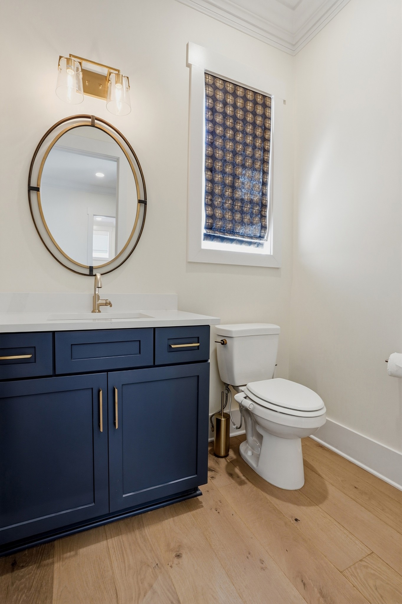 Bathroom with navy vanity, brass-framed oval mirror, patterned blue shade, and wood flooring.