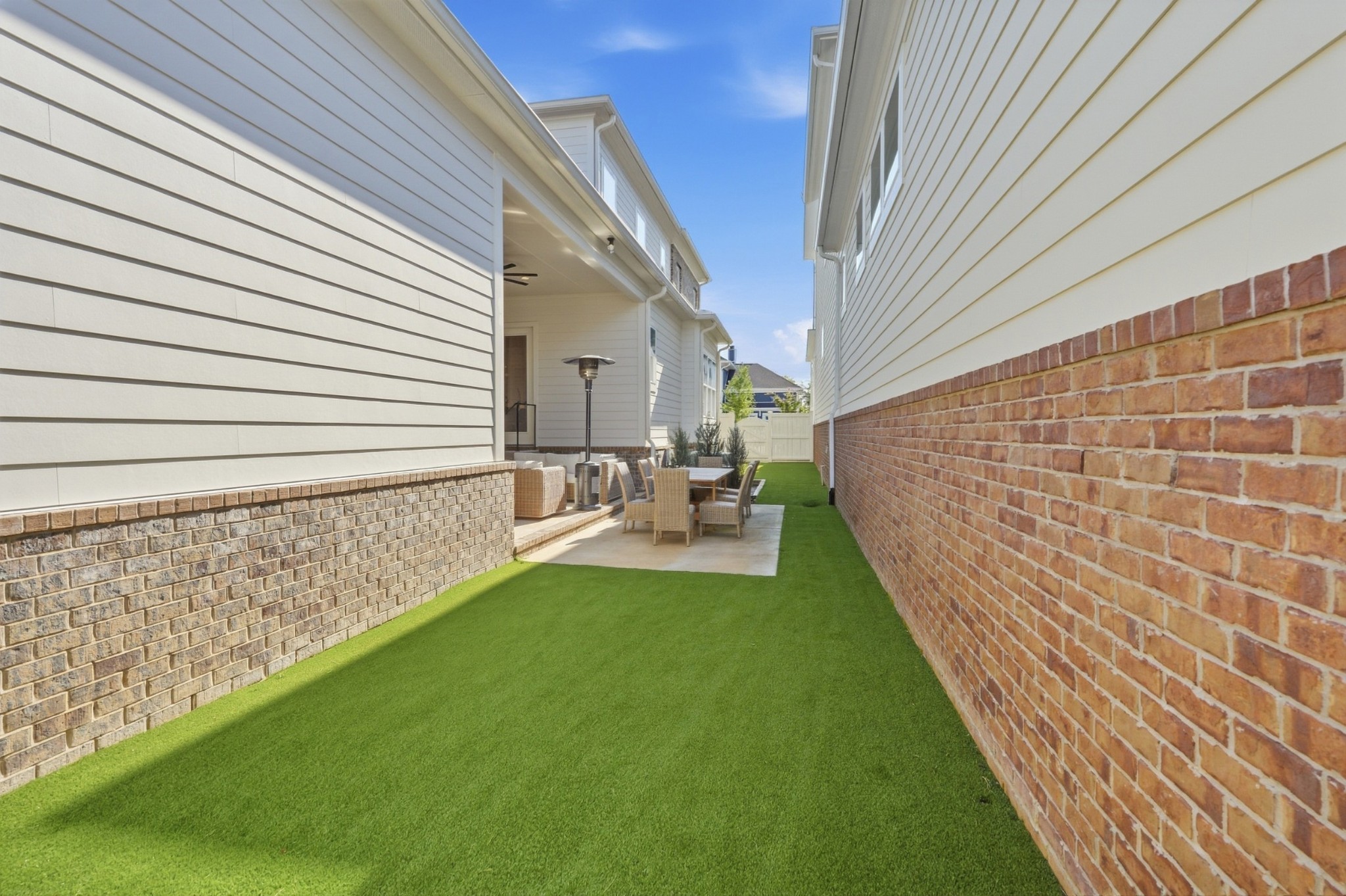 Narrow side yard with green lawn leading to a brick patio and outdoor dining set beneath a covered porch.