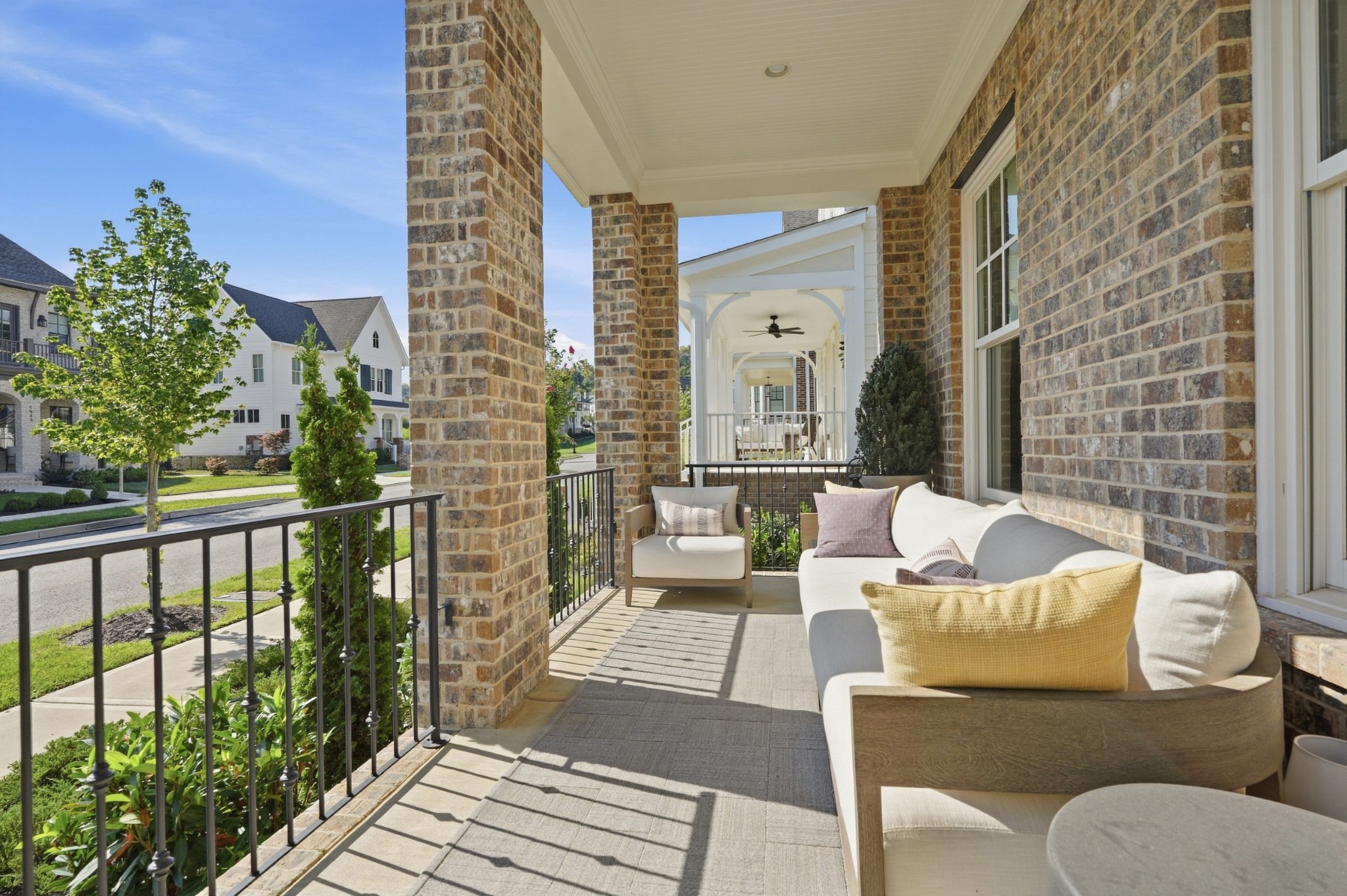 Covered brick porch with white outdoor seating, black railings, and a view of neighboring homes.