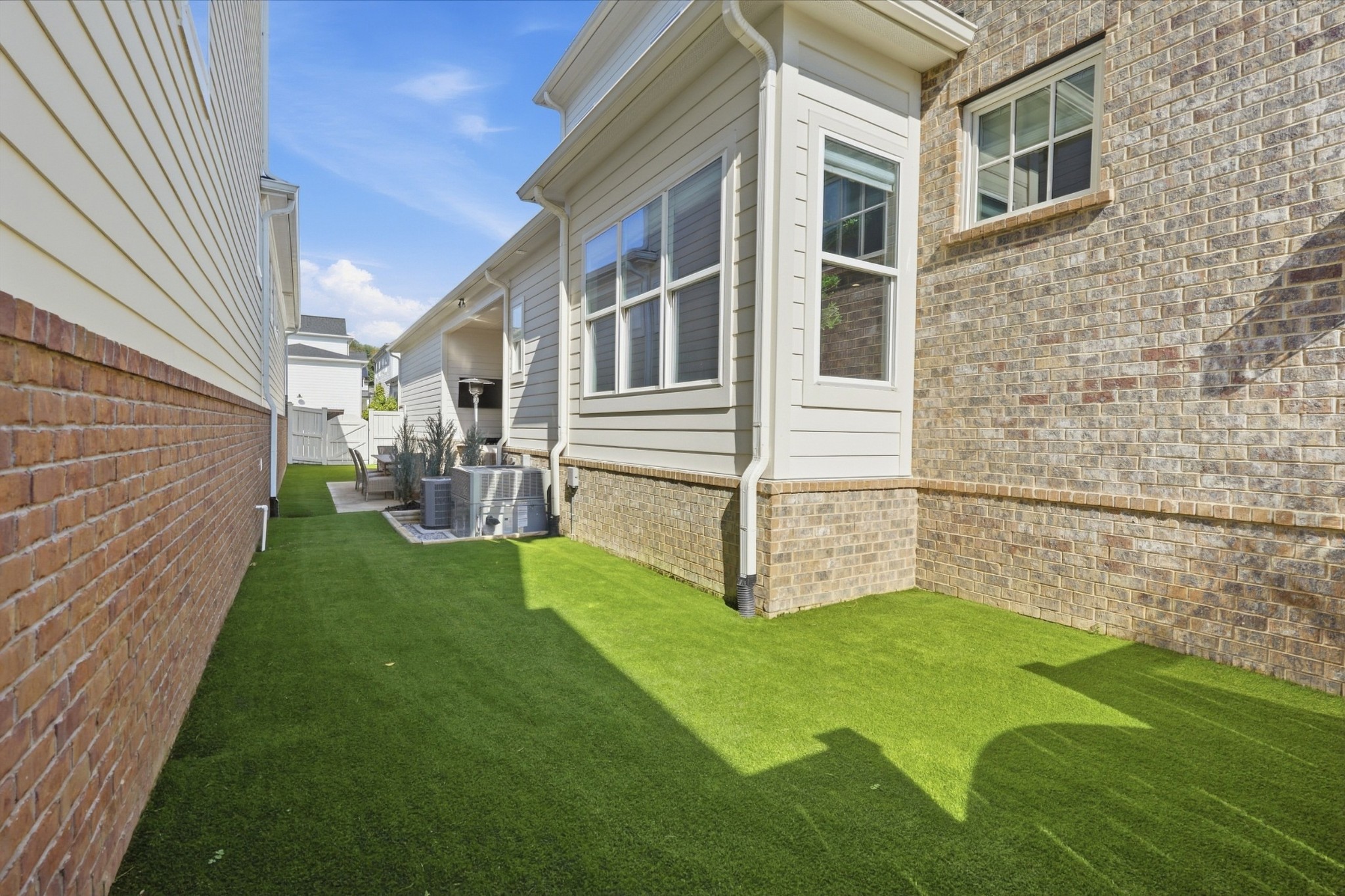 Side yard with green lawn beside a brick-and-siding home, with patio chairs and HVAC units.