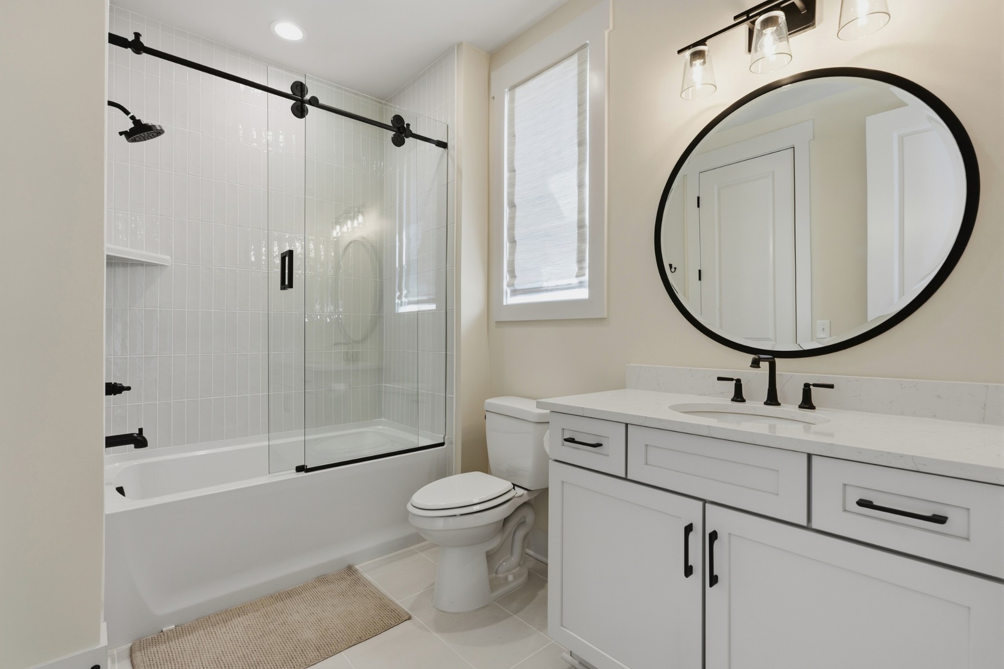Bathroom with a glass-enclosed tiled tub-shower, white vanity, and large round black-framed mirror.