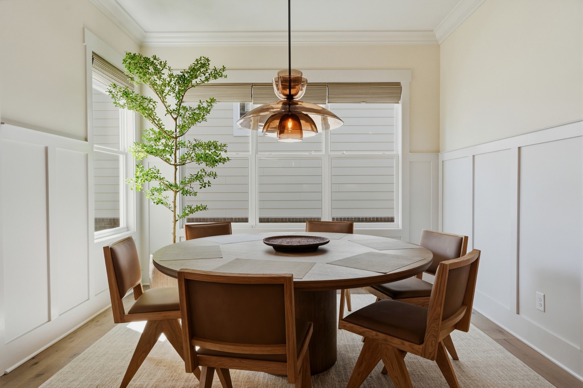 Dining room with a round wood table, brown chairs, large windows, and an amber glass pendant light.