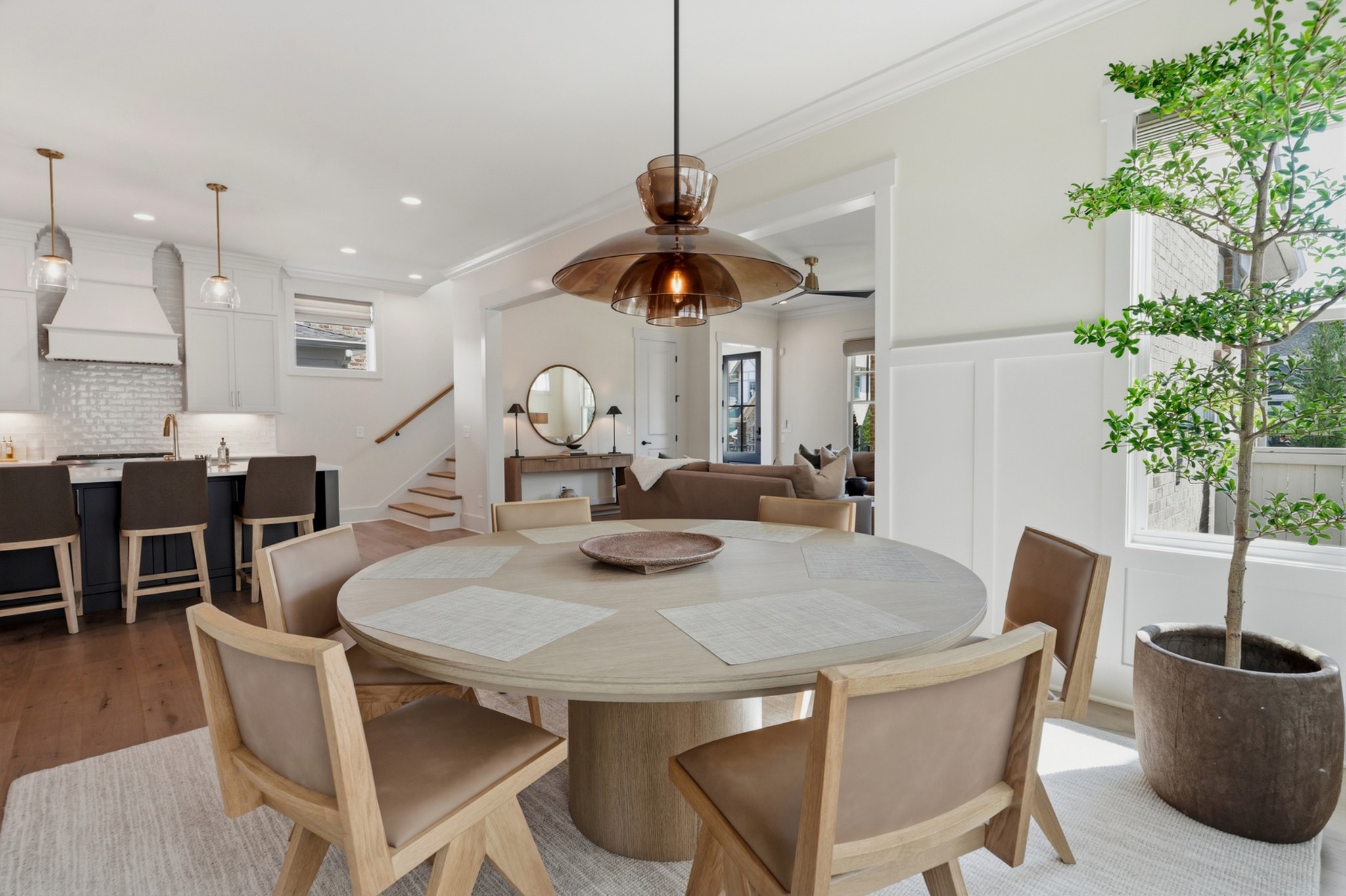 Open-plan dining room with a round wood table, smoked-glass pendant, white kitchen, and potted tree by the windows.