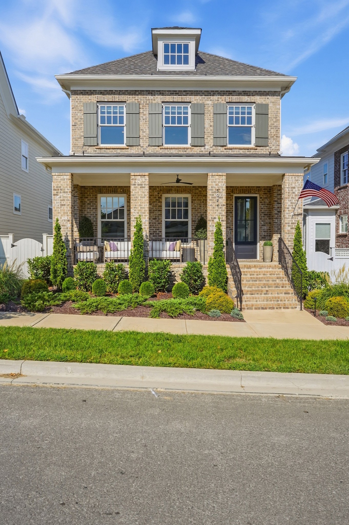 Two-story brick house with a furnished covered porch, landscaped front garden, and American flag.