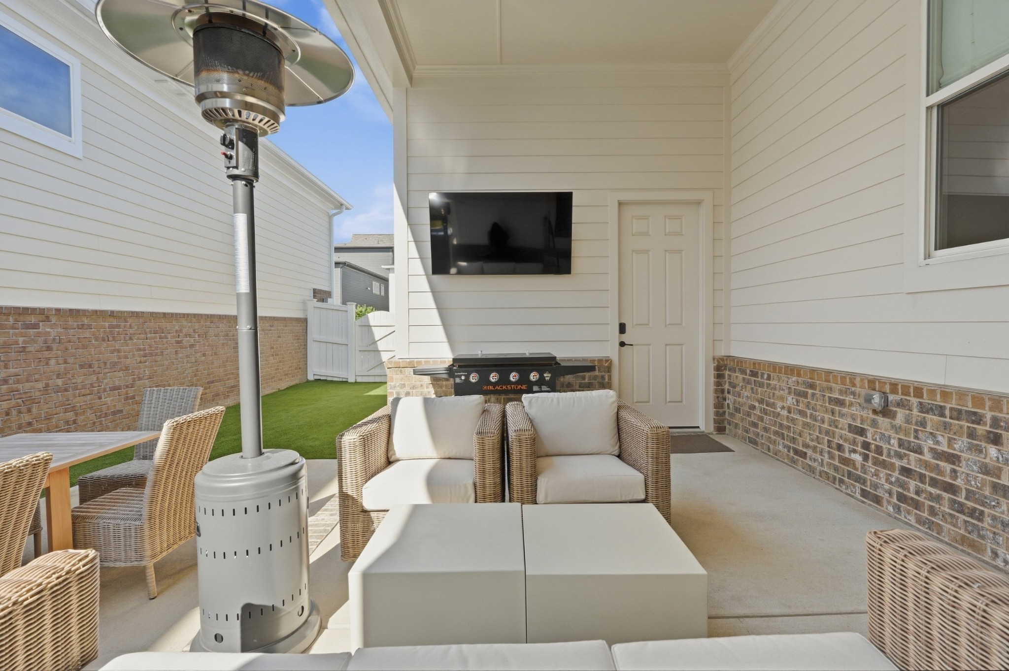 Covered patio with wicker seating, a patio heater, wall-mounted TV, and grill beside a small fenced turf yard.