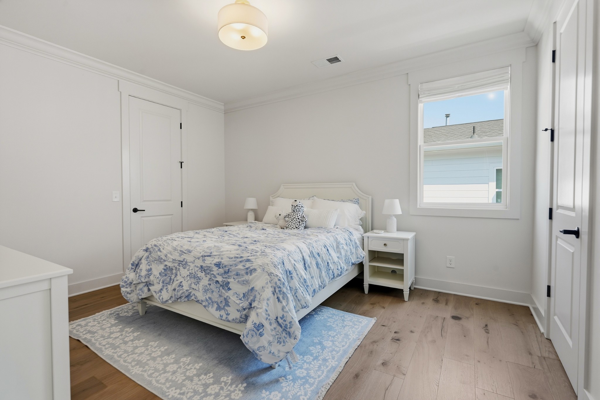 Staged bedroom with a white bed, blue floral bedding, bedside tables, and a window overlooking a neighboring house.
