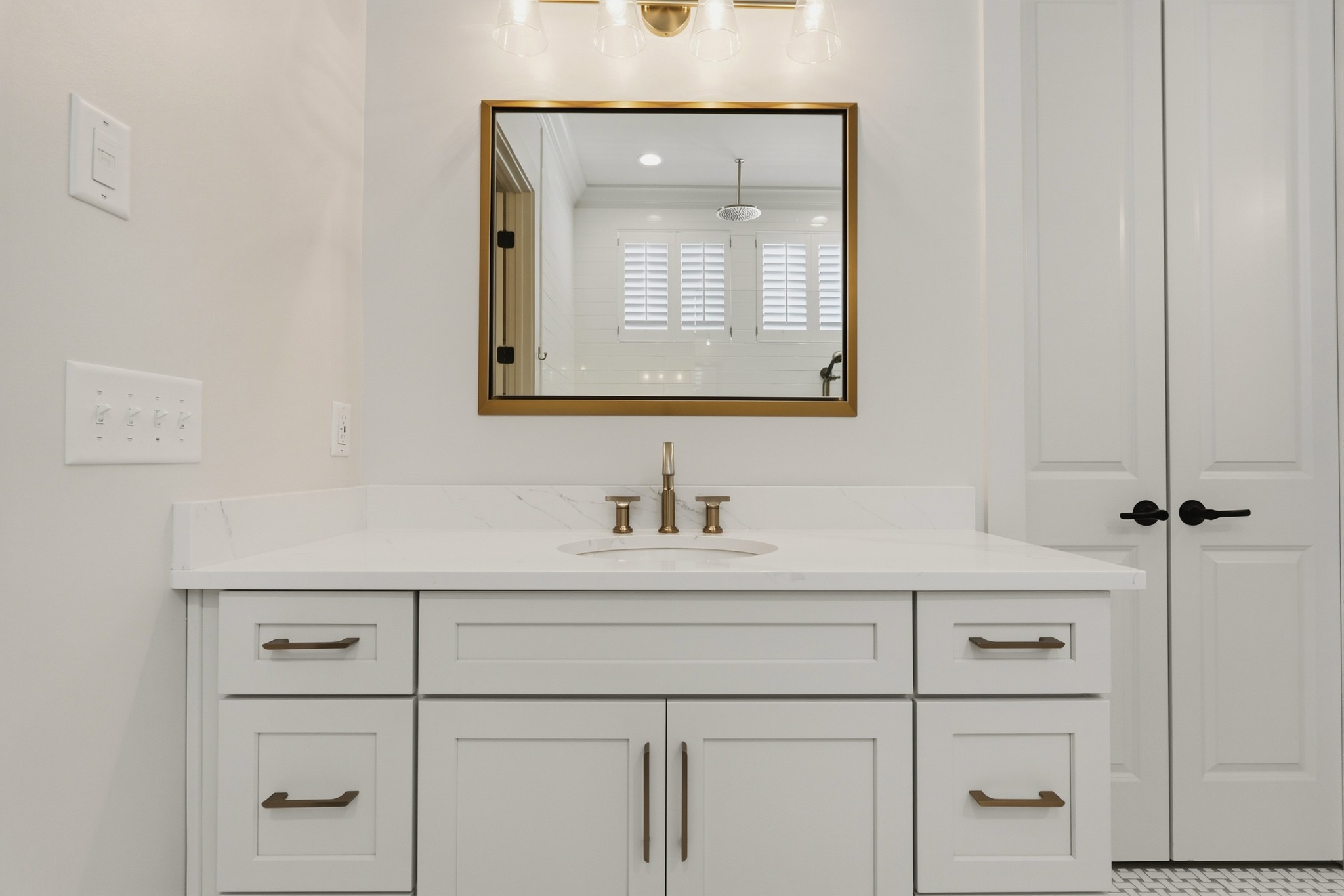 Bathroom vanity with brass fixtures and a large mirror reflecting a shower with shuttered windows.