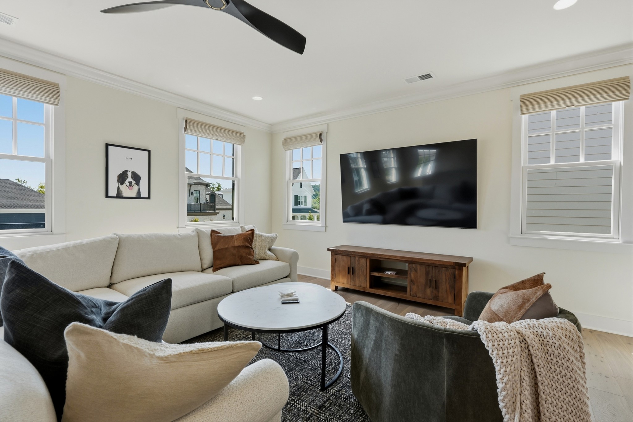 Furnished living room with a cream sofa, round coffee table, wall-mounted TV, and bright windows.