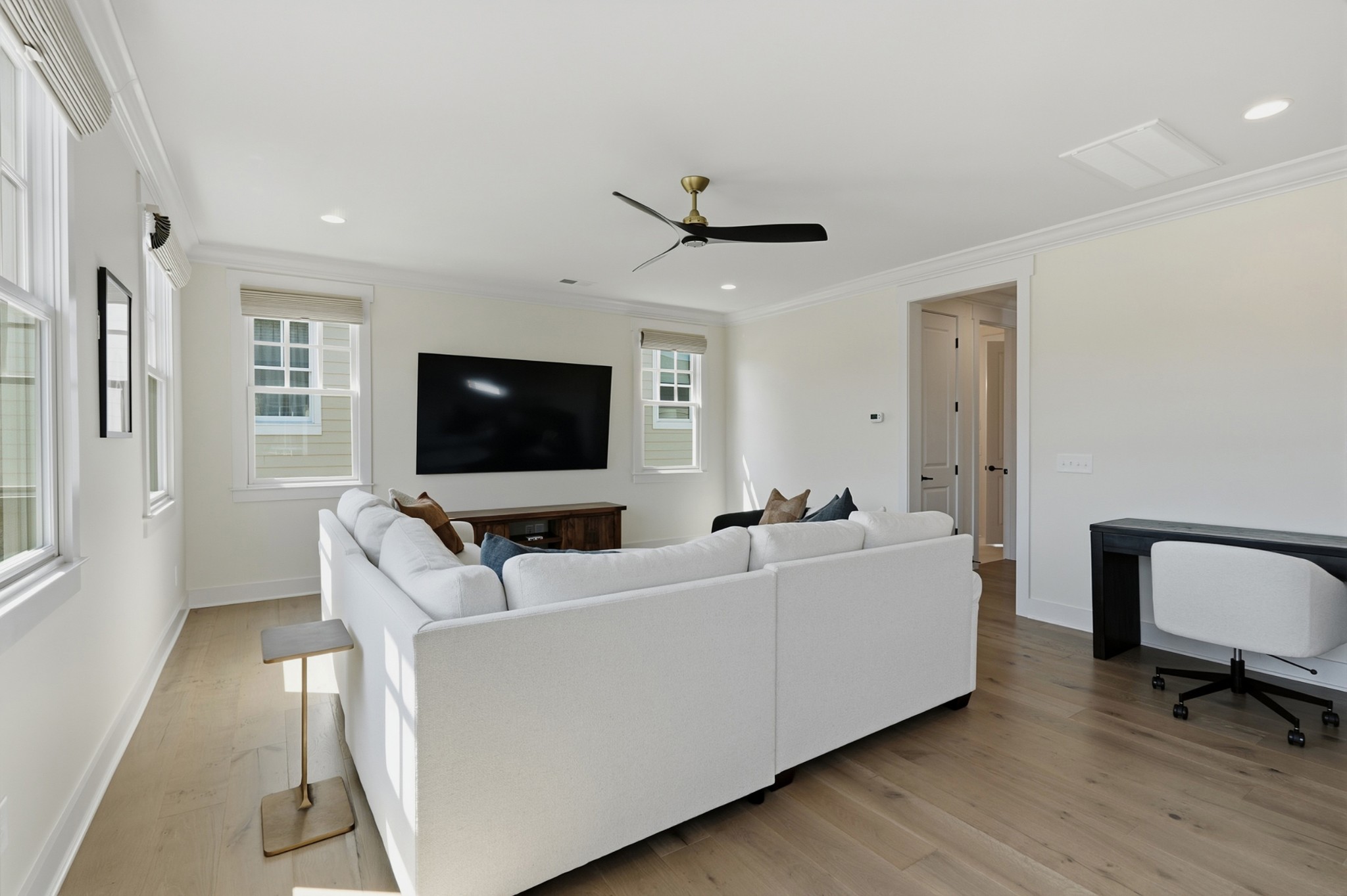 Bright living room with a large white sectional sofa, wall-mounted TV, and several sunlit windows.