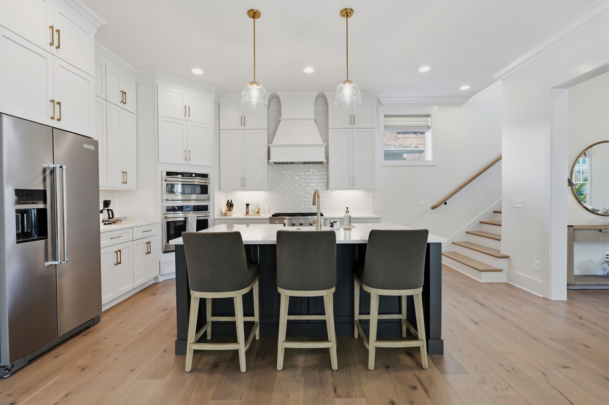 Modern white kitchen with a black island, three stools, stainless steel appliances, and a staircase.