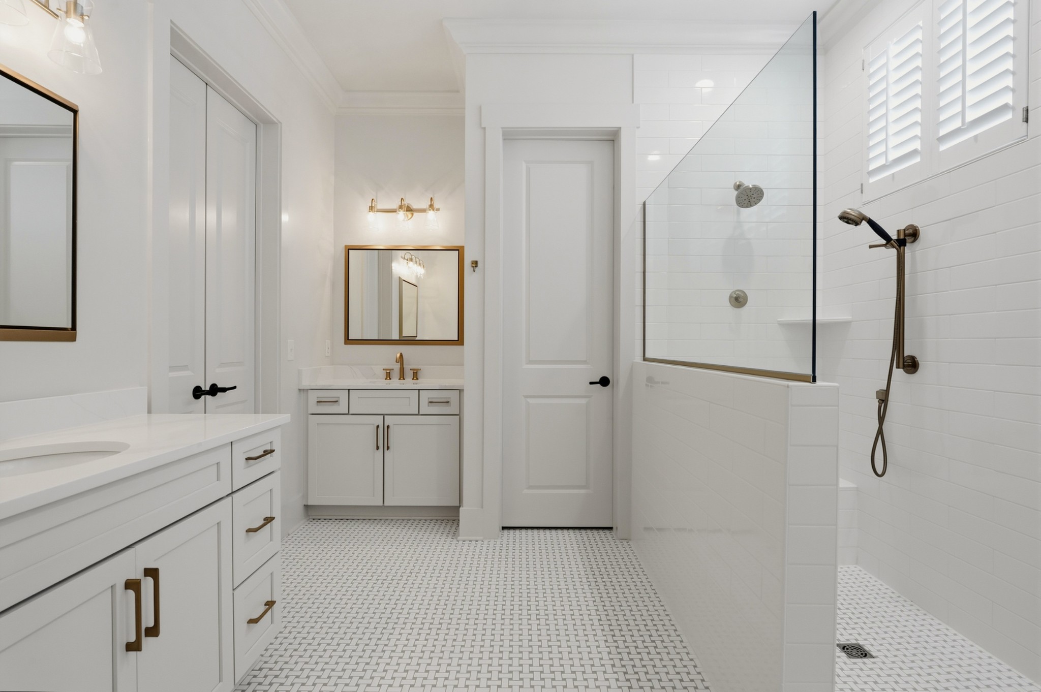 White bathroom with a double vanity, brass fixtures, patterned tile floor, and glass-enclosed shower.