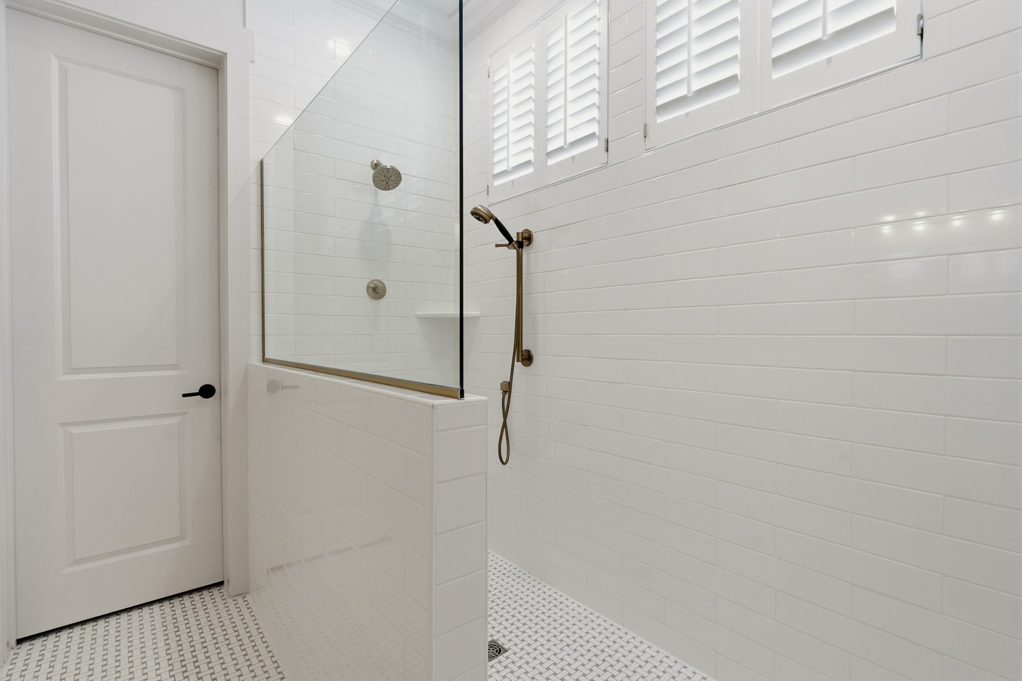 White-tiled walk-in shower with glass panel, brass fixtures, shuttered windows, and mosaic floor.