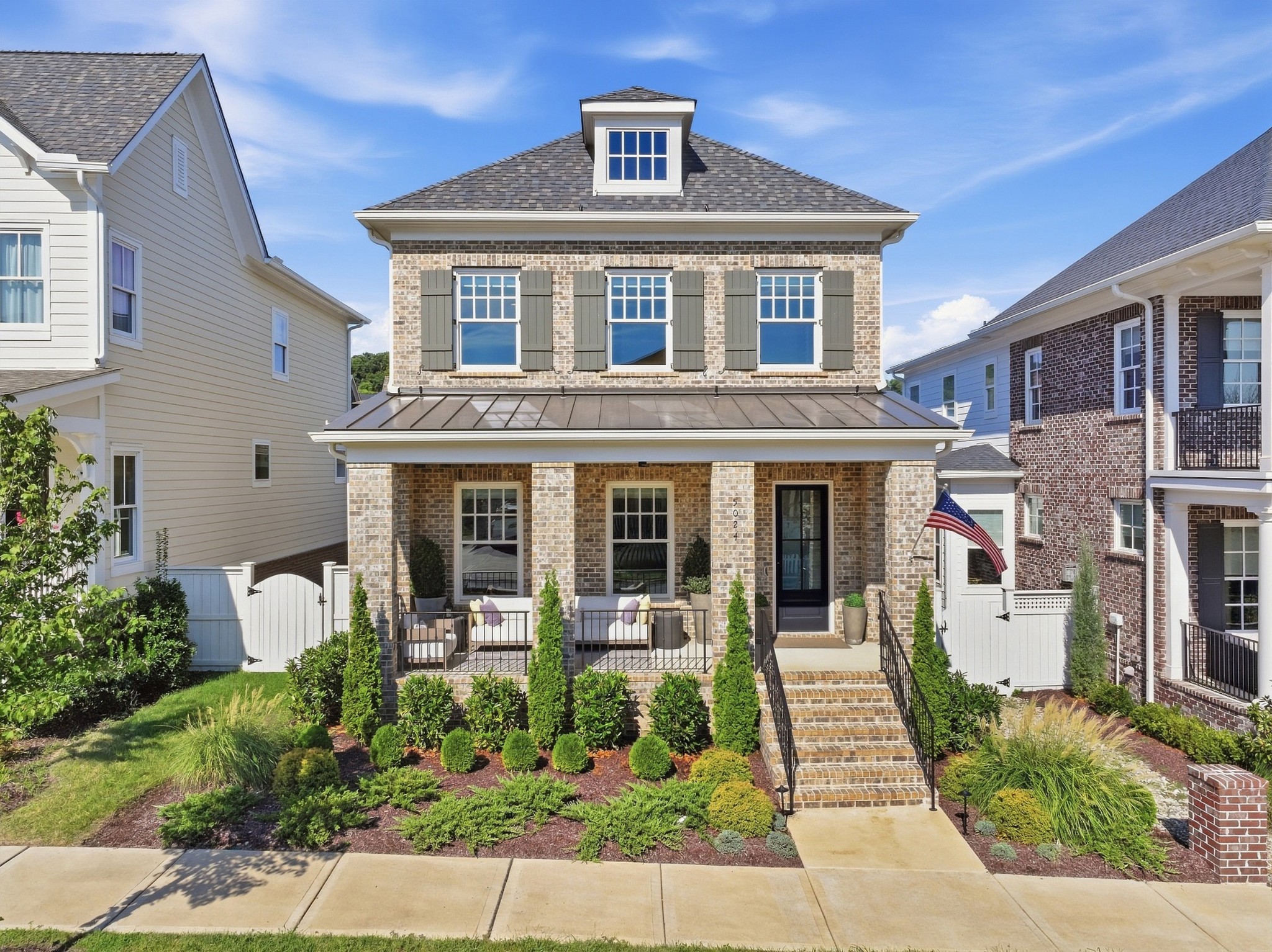 Two-story brick home with a covered porch, landscaped front garden, and American flag.