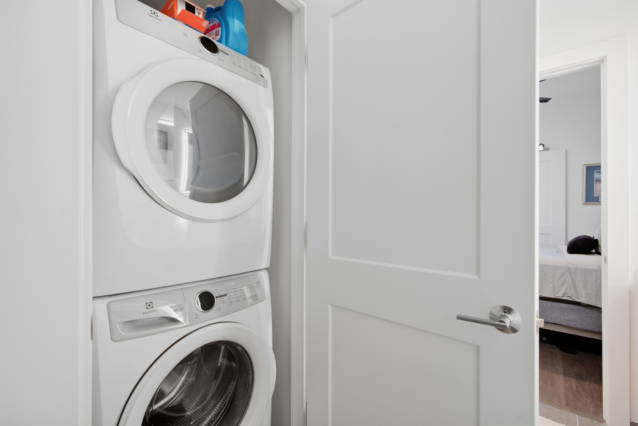Laundry closet with stacked white washer and dryer beside an open doorway to a bedroom.
