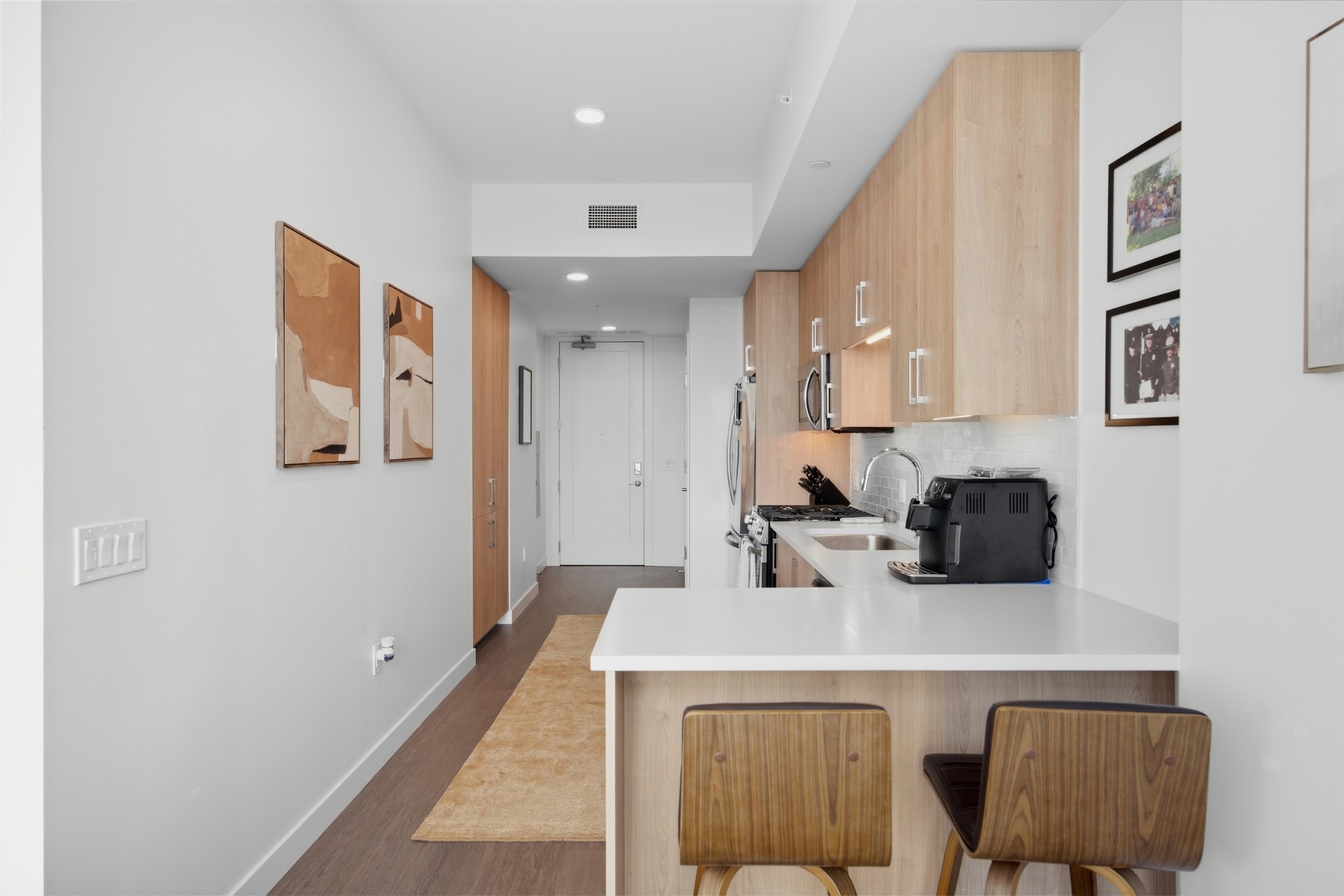 Modern condo kitchen with light wood cabinetry, white counters, and a breakfast bar with two stools.