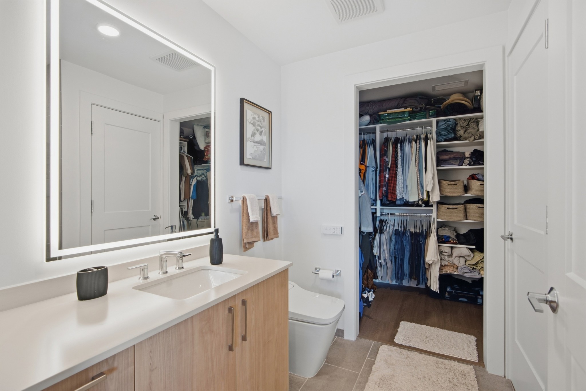 Bathroom with a light wood vanity and large mirror opening onto a walk-in closet filled with clothes.