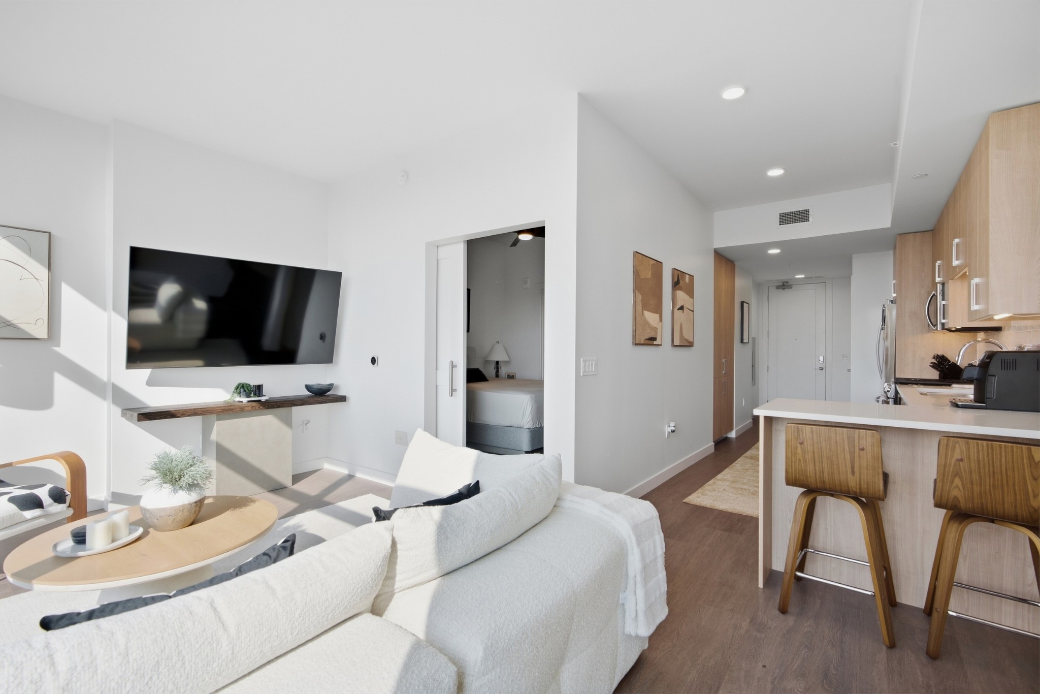Furnished condo living room with a white sofa, wall-mounted TV, and kitchen island with bar stools.