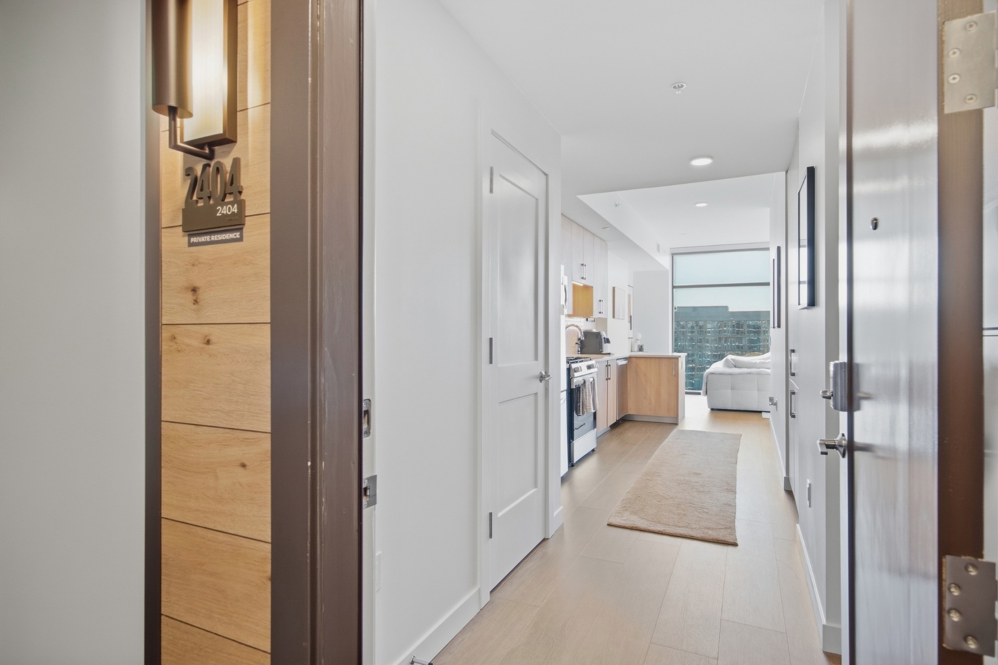 Condo entry hallway with light wood floors, compact kitchen, and living area beyond.