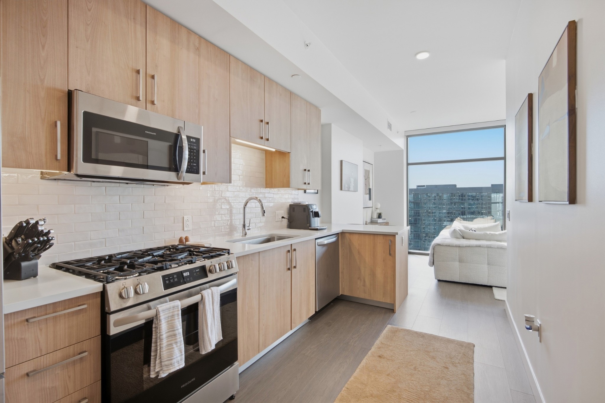 Condo kitchen with light wood cabinets, stainless appliances, and a large window beside the living area.