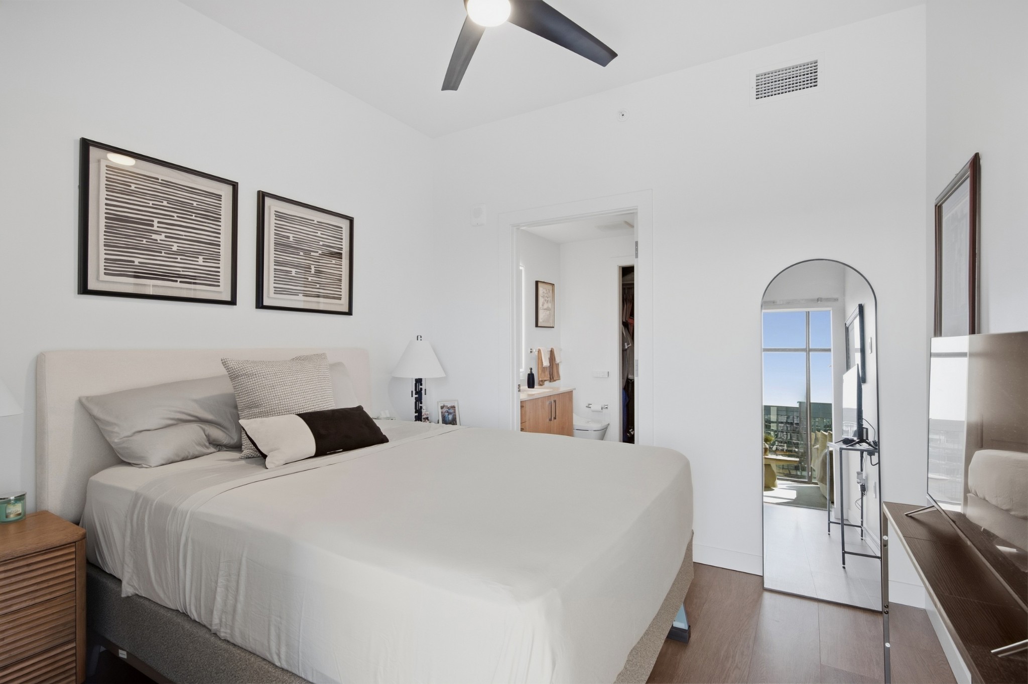 Furnished bedroom with a large bed and mirror reflecting a balcony and city view.