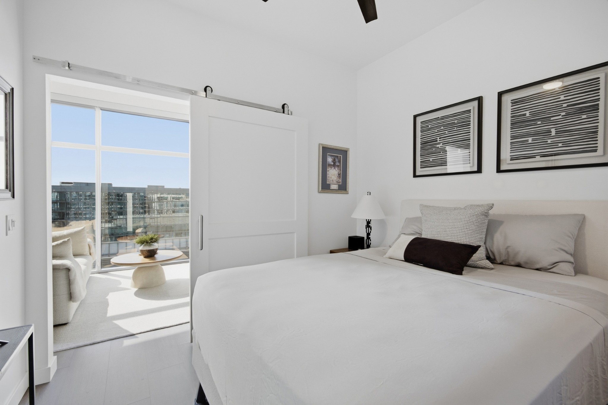 Bedroom with a large bed and barn door opening to a sunny balcony with city views.