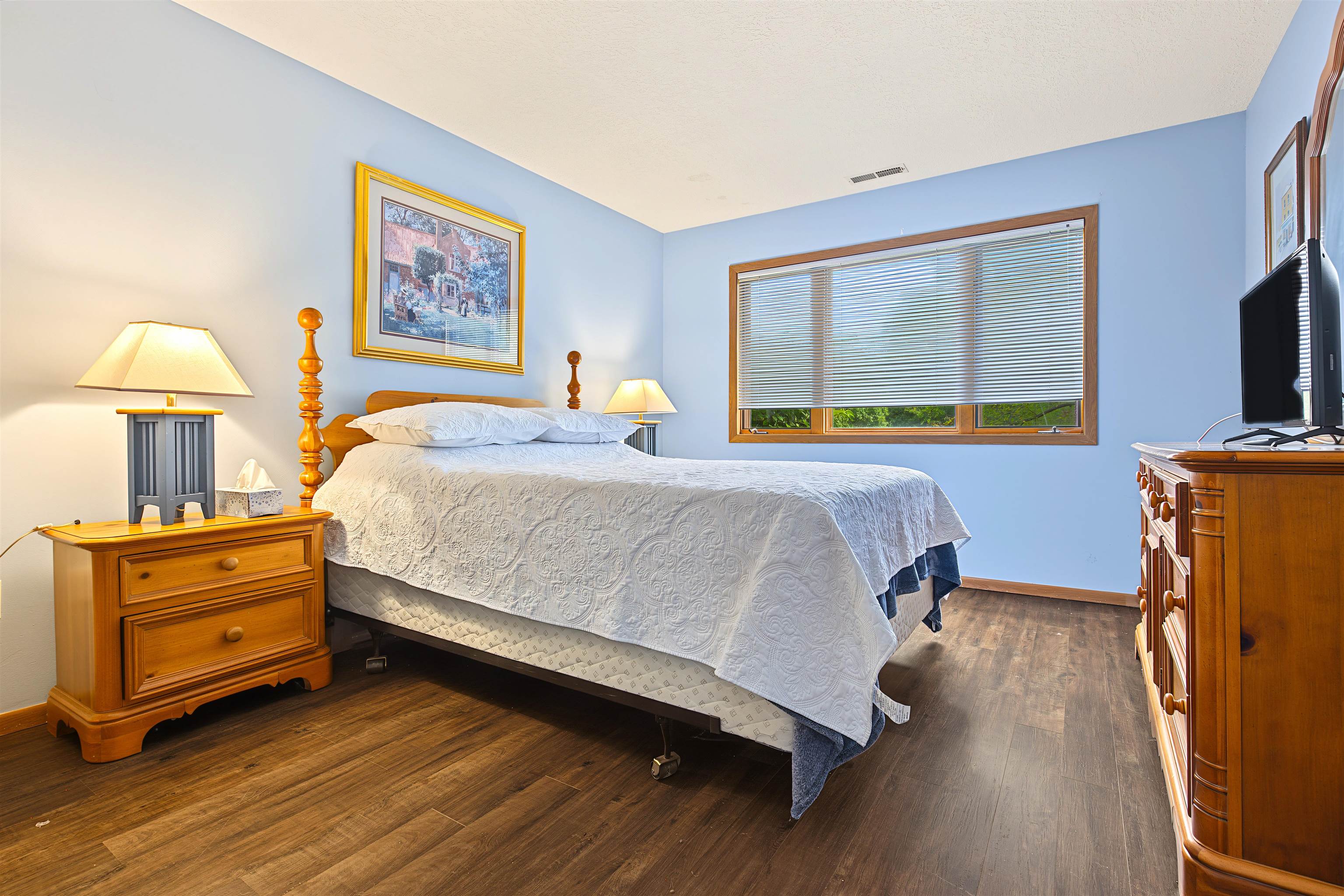 Furnished bedroom with a large white-quilted bed, blue walls, wooden furniture, and a wide window.
