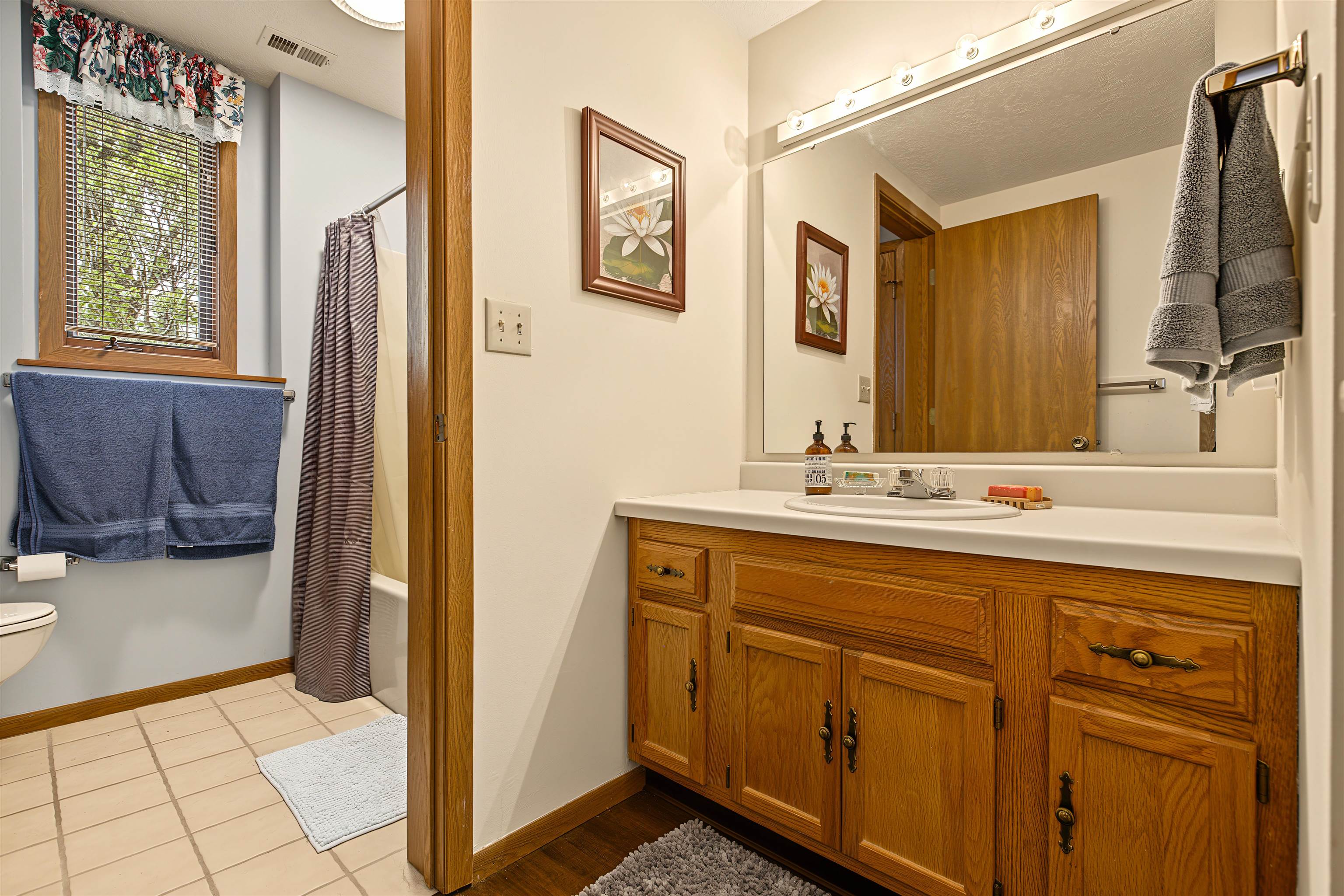 Bathroom with an oak vanity and large mirror, beside a tiled shower area and window.