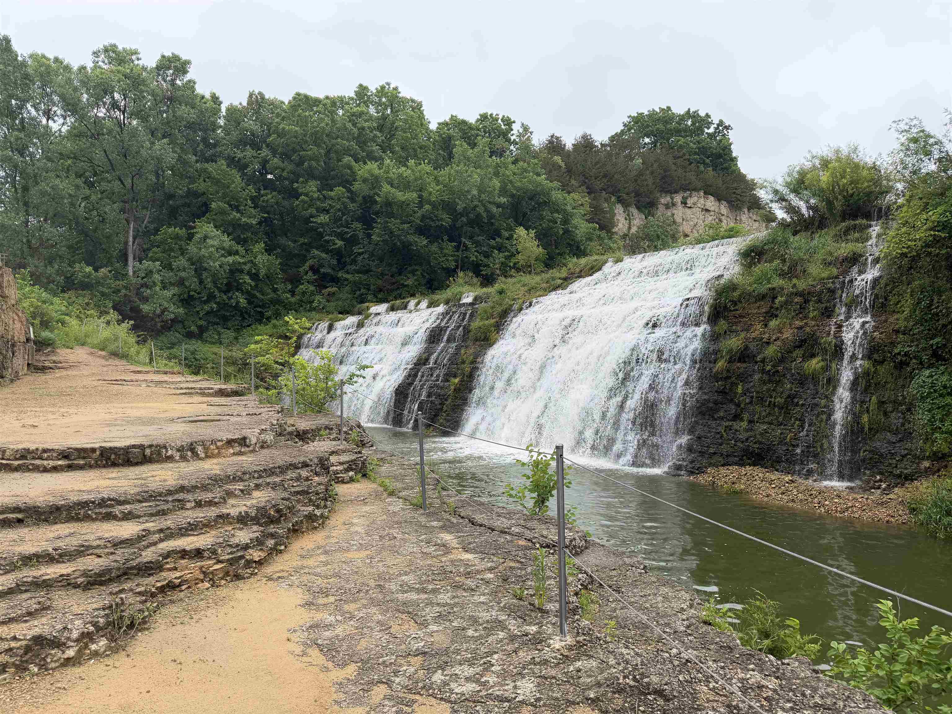 Waterfall beside a stone walkway, with wooded banks and rope railings.
