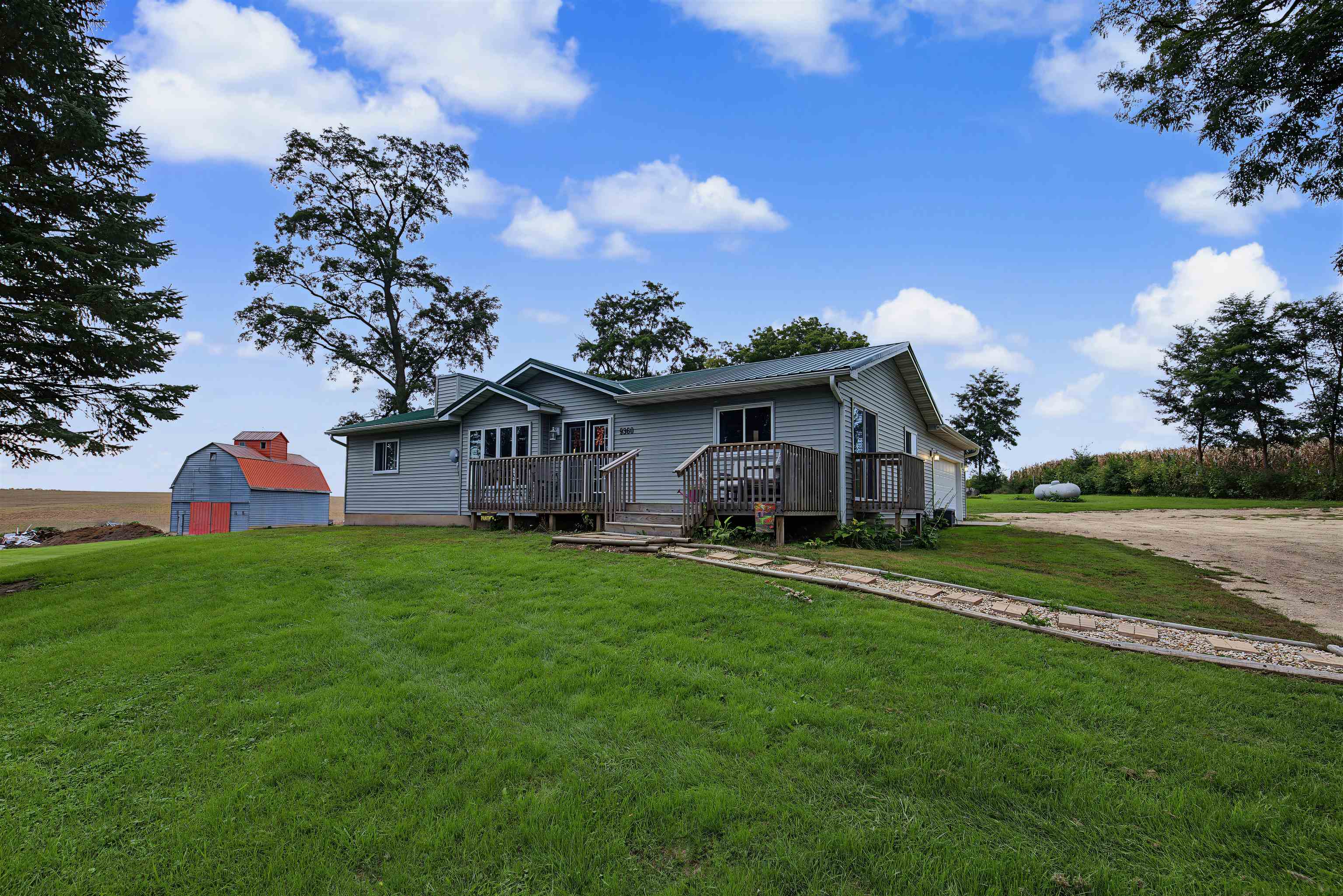 Gray rural home with a raised deck and metal roof beside a blue-and-red barn and broad lawn.