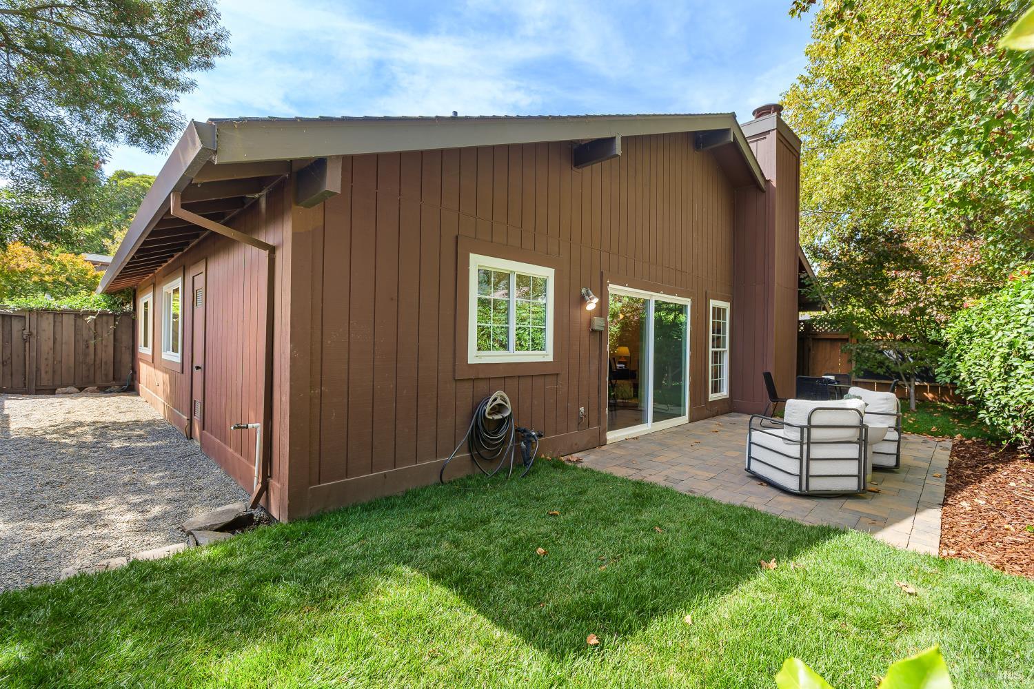 Brown-sided home's rear exterior with a paved patio, outdoor seating, and surrounding lawn and trees.