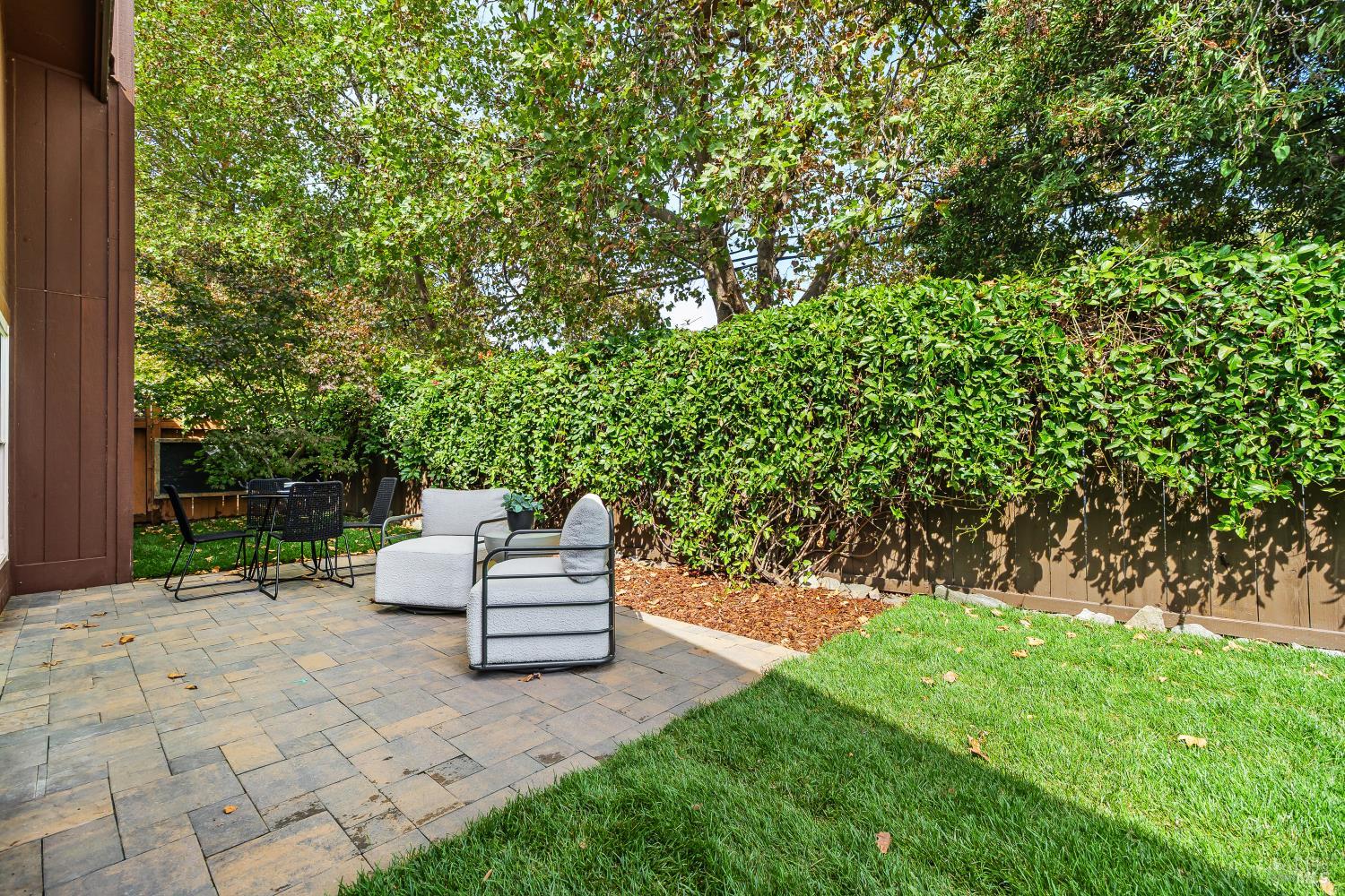 Backyard patio with stone pavers, outdoor seating, a green lawn, and a tall hedge.