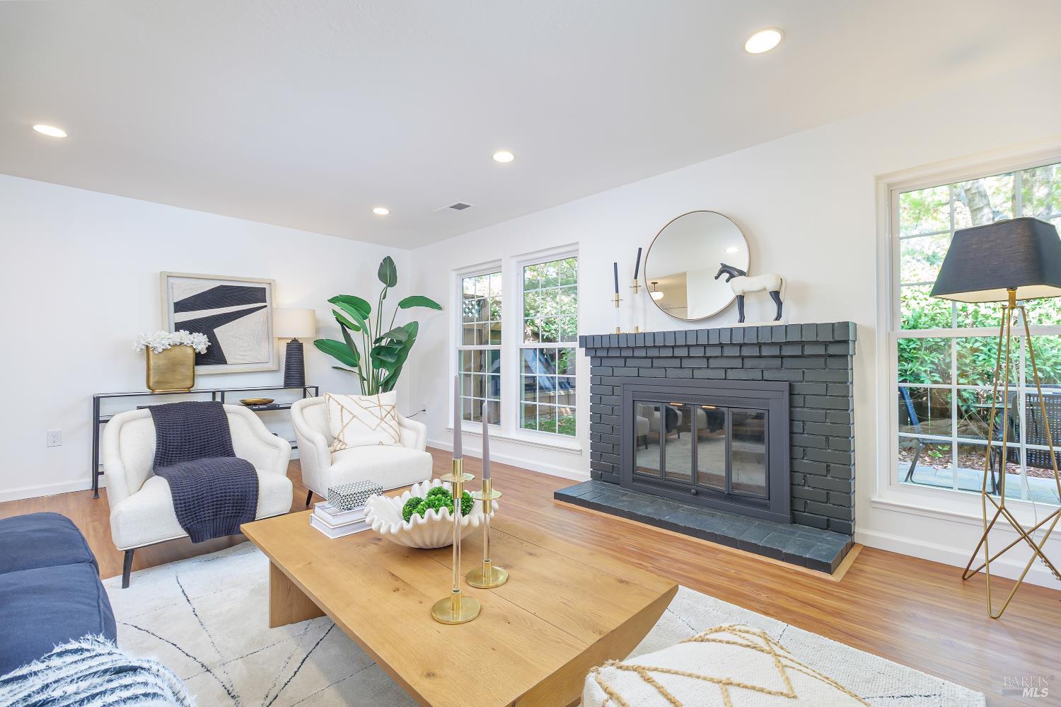 Staged living room with a dark brick fireplace, white armchairs, and large windows overlooking greenery.