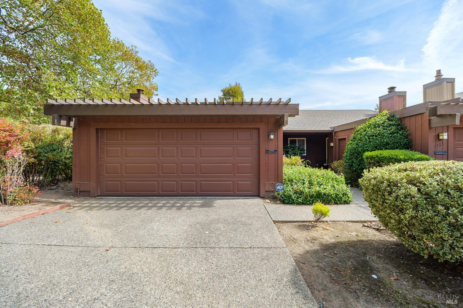 Brown home exterior with a wide double garage, pergola-style roof beams, and landscaped shrubs.