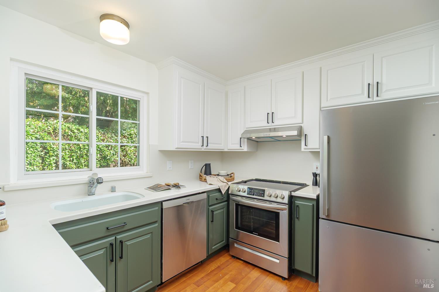 Kitchen with sage-green lower cabinets, white cabinetry, and stainless steel appliances beside a large window.