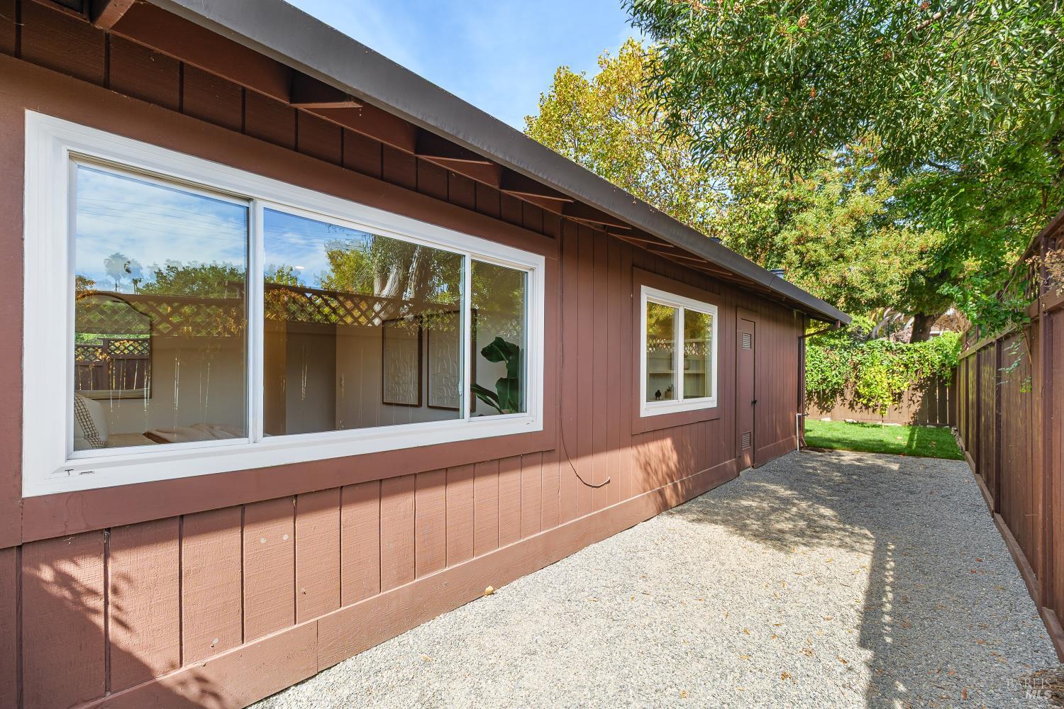 Brown home exterior with white-trimmed windows beside a gravel path and fenced, tree-shaded yard.