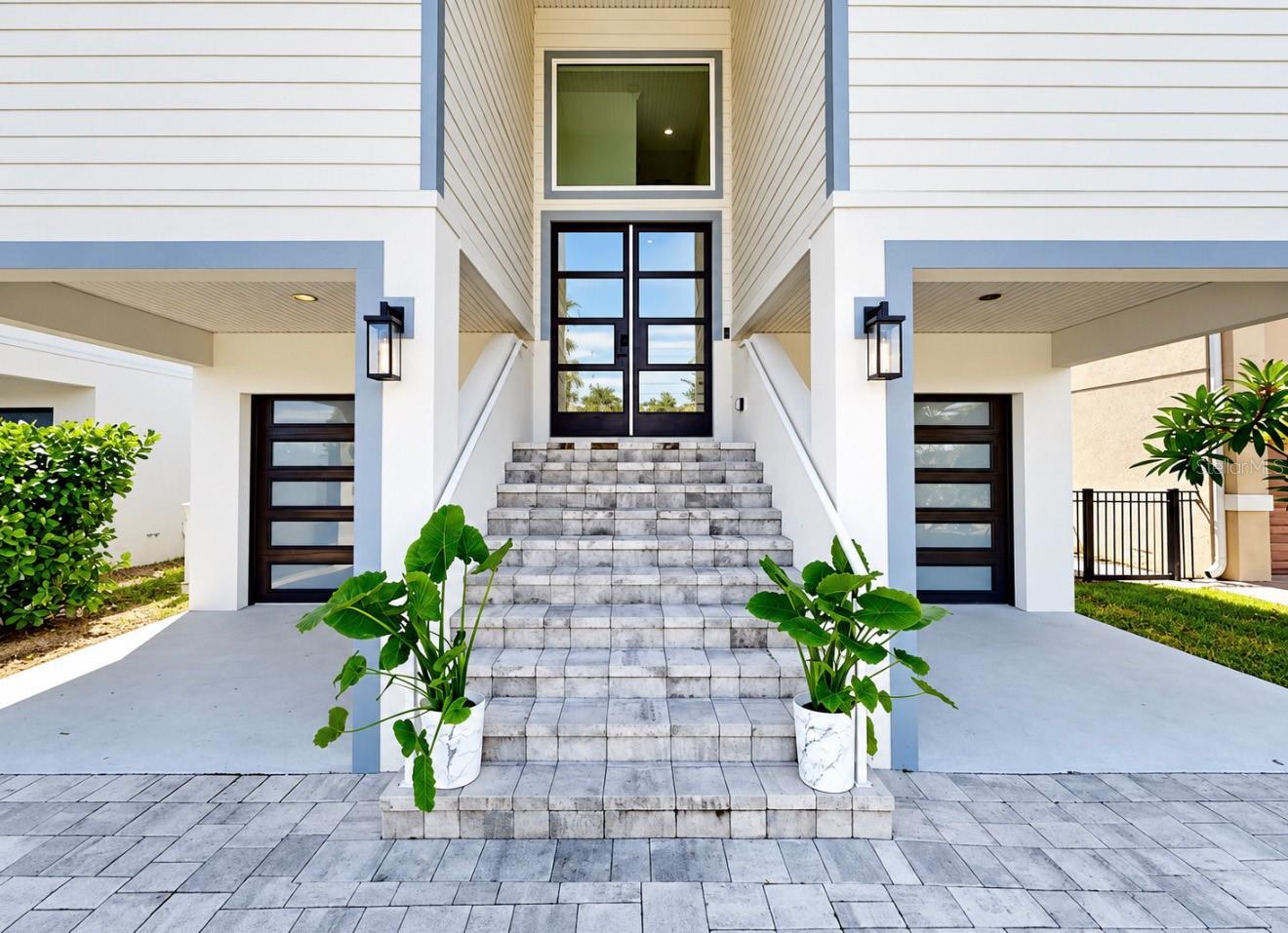 Home entrance with broad tiled steps, black glass doors, blue trim, and potted tropical plants.