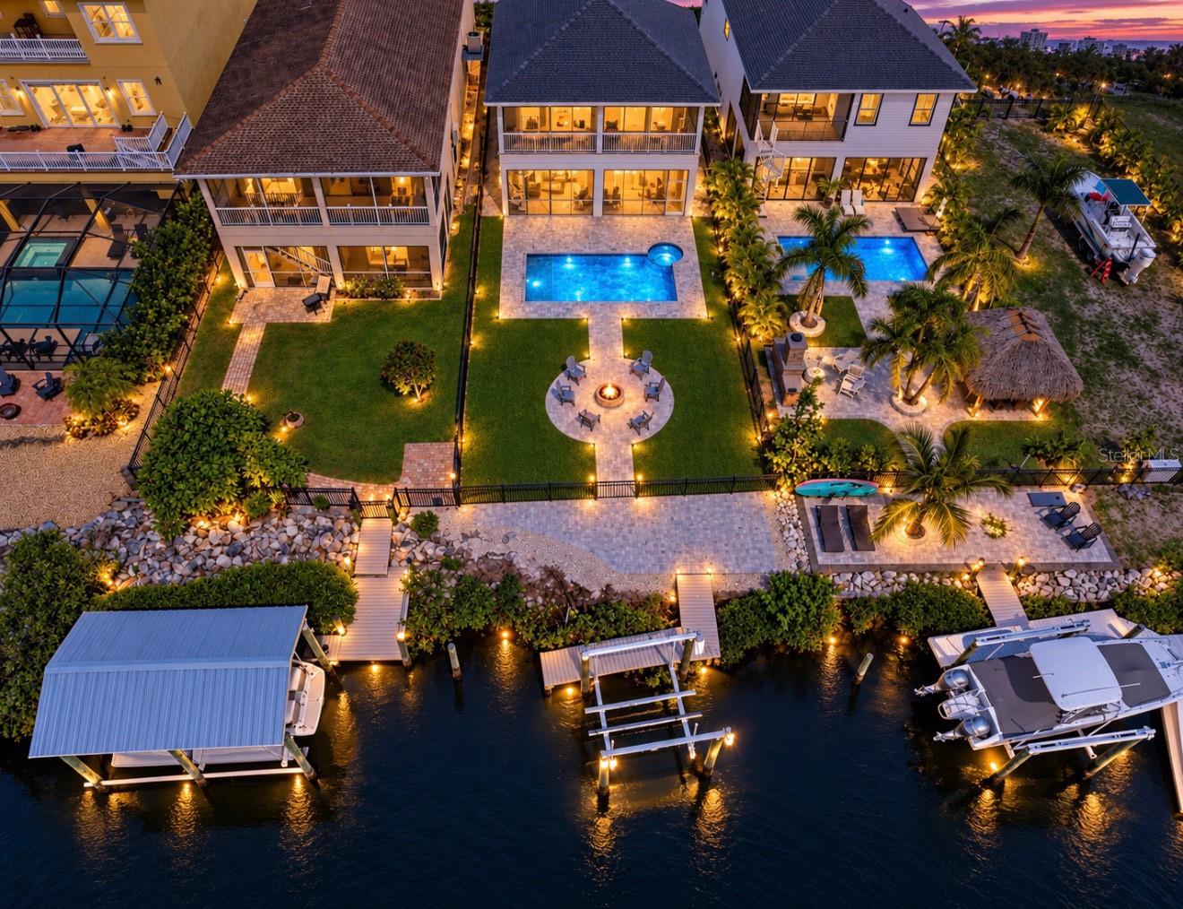 Waterfront home with a lit pool, landscaped yard, fire pit, and private dock at dusk.