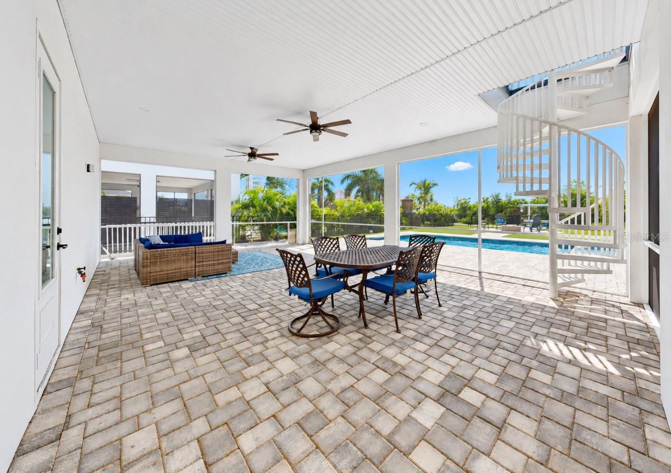 Covered patio with outdoor dining and wicker seating beside a pool, with a white spiral staircase.