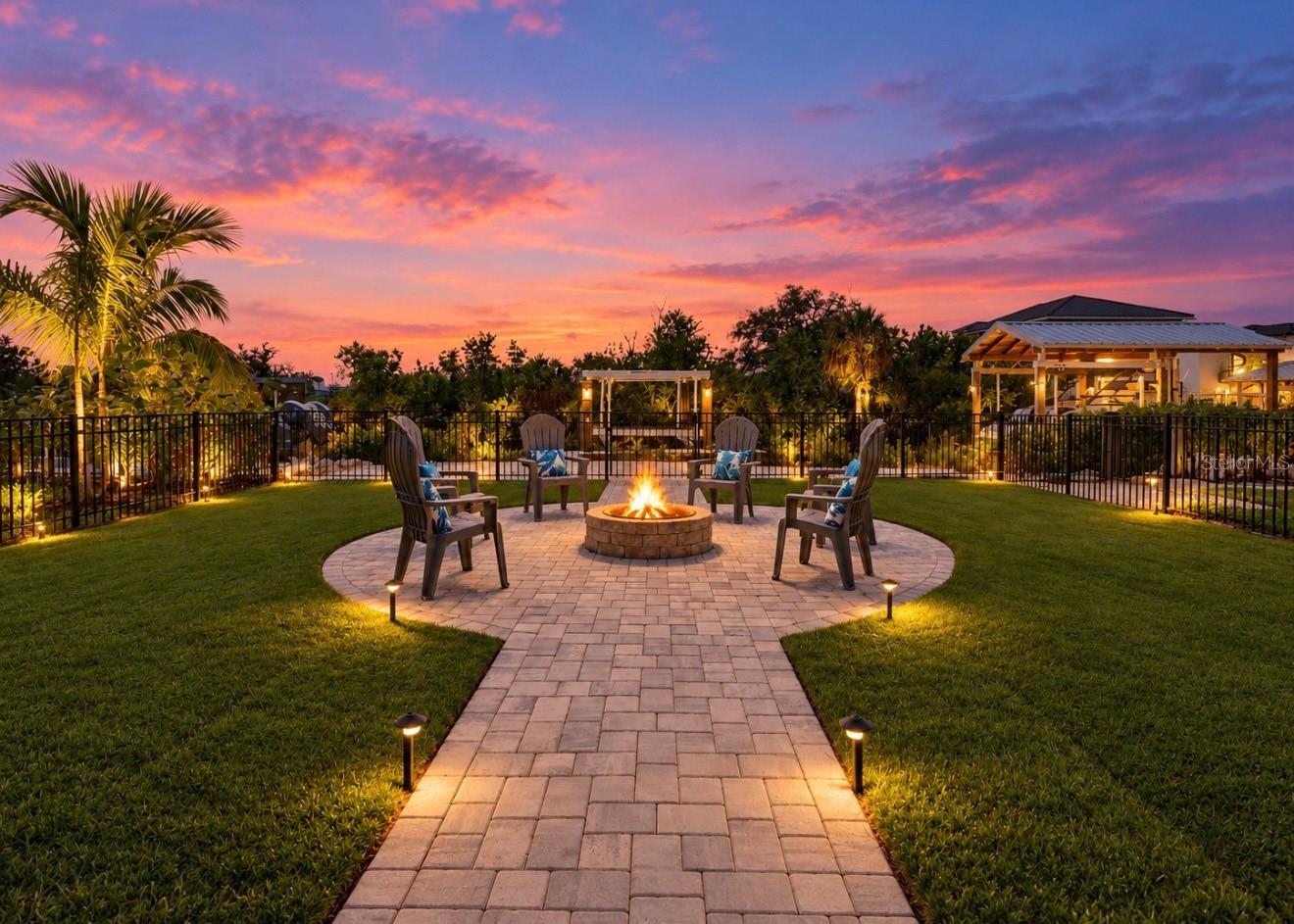 Backyard patio with a circular fire pit seating area and tropical landscaping at sunset.