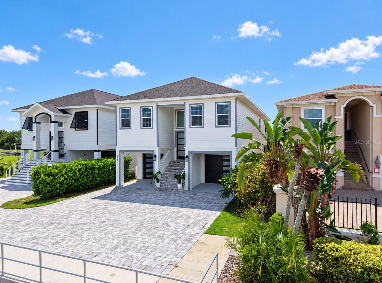 White two-story home with an elevated entry, garage, paver driveway, and tropical landscaping.