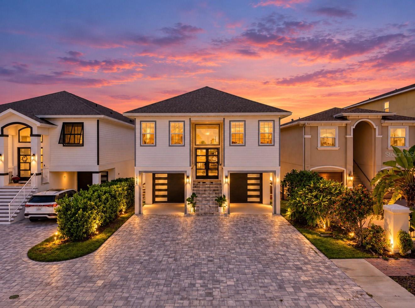 Two-story coastal home with twin garage doors and a paver driveway at sunset.